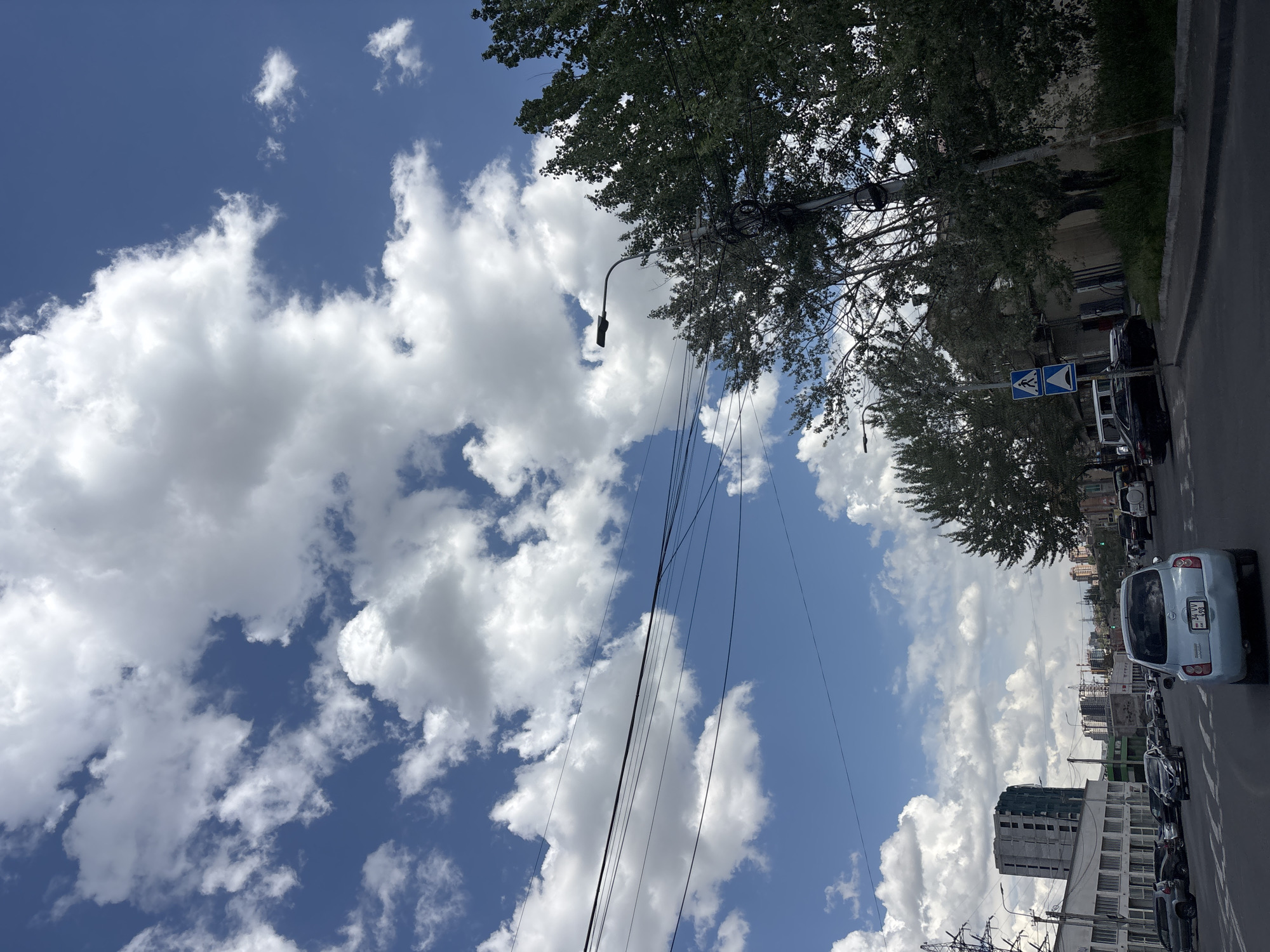 The image shows a street scene in Yerevan, Armenia, under a partly cloudy sky. The sky is blue with numerous white cumulus clouds. On the right side of the image, a road with parked cars is visible. Among the vehicles is a light blue car with Armenian license plate. Pedestrian crossing signs are seen near the cars. Above the cars, trees line the street. Power lines stretch across the sky, appearing tangled. In the background, a building under construction is visible, along with other buildings that compose the city landscape.