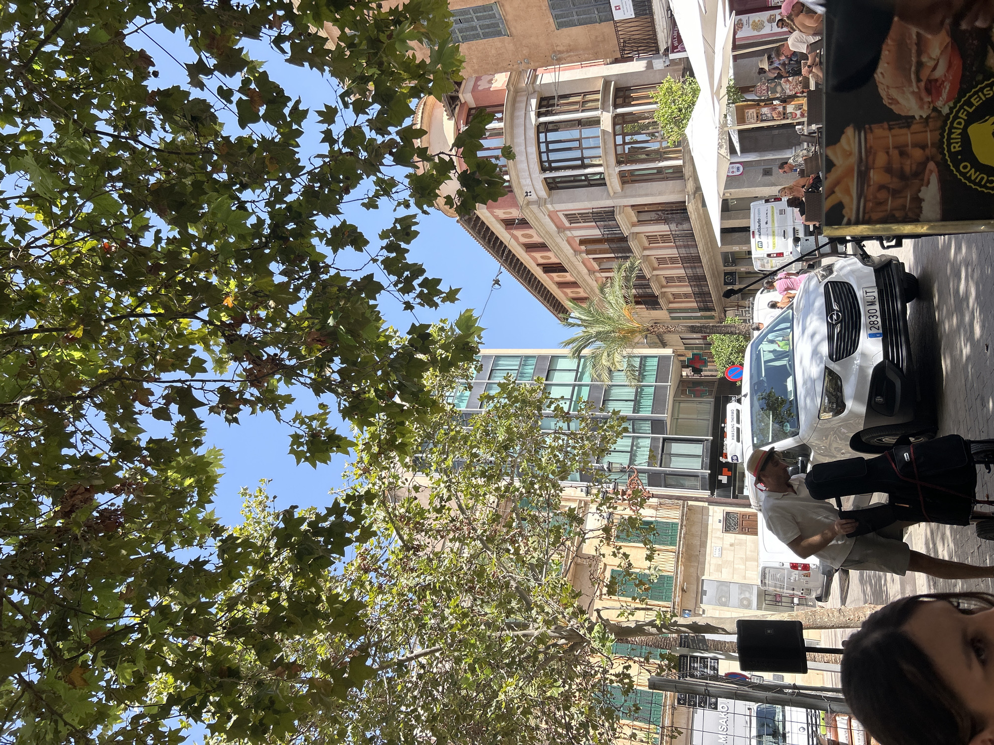 This image captures a street scene in Manacor, Spain, on a sunny day, with a focus on a white Opel vehicle parked on the street. The vehicle, a hatchback with a license plate reading "2830 MJT," is positioned in the foreground, partially obscured by a person wearing a straw hat and light-colored clothing, who is interacting with a black rolling case.
The background features a street lined with buildings of varying architectural styles. To the left, a modern building with a glass facade stands tall. Adjacent to it is a building with traditional elements, characterized by light-colored walls and green shutters. Further to the right, a pink and beige building with ornate balconies and rounded bay windows suggests a more classic European design. A palm tree is visible near this building, adding a tropical touch to the urban landscape.
Several white umbrellas from outdoor seating areas of what appear to be cafes or restaurants are spread across the scene. People are seated at tables under these umbrellas, suggesting a bustling atmosphere. Overhead, the branches and leaves of a large tree partially obscure the blue sky, casting dappled shadows on the street. The overall impression is a lively European city street during daylight hours.