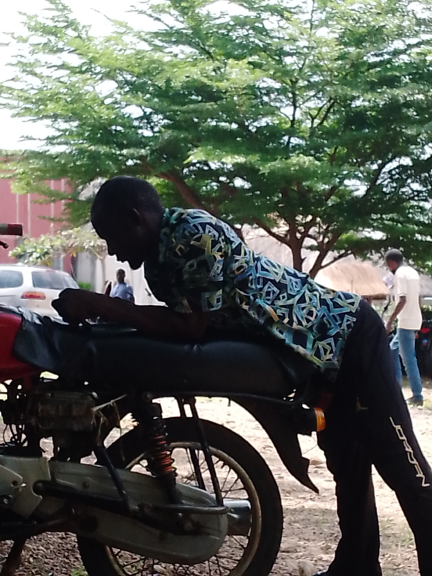 The image depicts two individuals in what appears to be an outdoor setting in Akwanga, Nigeria. In the foreground, a young man is leaning over a motorcycle, his body angled downwards. He is wearing a patterned short-sleeved shirt with a design of geometric shapes in blue, green, and white, and dark pants. His hands are clasped in front of him on the motorcycle's seat. The motorcycle is dark-colored with red accents on the front fender and a visible shock absorber.
In the background, another person, a man wearing a white t-shirt and blue jeans, is visible standing. Behind him, there is a white car, a building with a red facade, and lush green trees. The presence of trees and the natural lighting suggest it is daytime, likely with pleasant weather given the outdoor activity. The scene appears to be casual, possibly a roadside or a gathering spot. The word "JORDAN" is visible vertically printed on the dark pants of the man in the foreground.