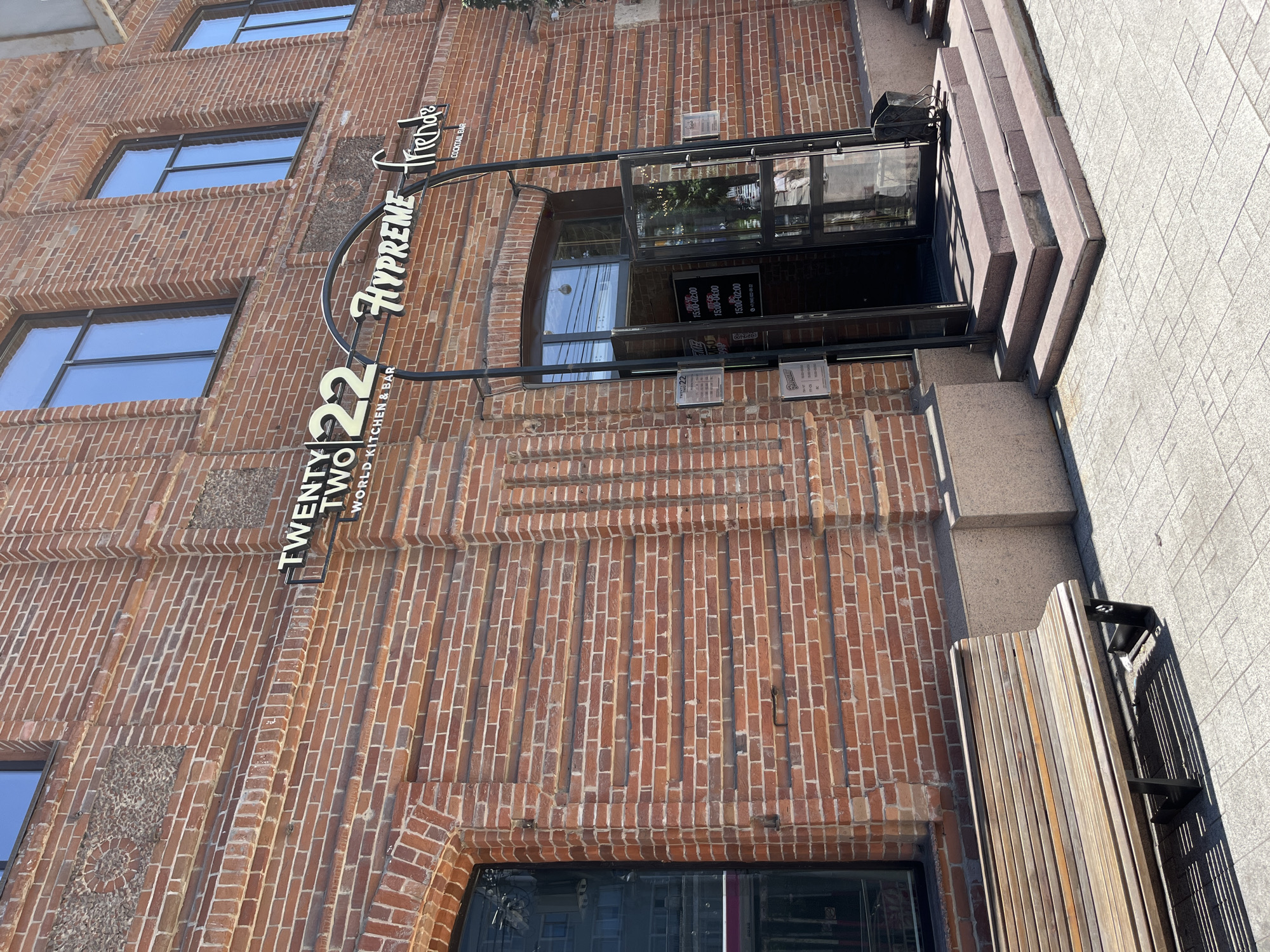The image shows the exterior of a brick building with a sign that reads "TWENTY TWO 22 HYPREME friends COCKTAIL BAR". The entrance to the establishment is a set of stairs leading to glass doors. To the left of the entrance, there is a wooden bench on a tiled sidewalk. The building appears to be in an urban setting, with windows visible on the upper floors. The lighting suggests it is daytime. There are no people clearly visible in the image.