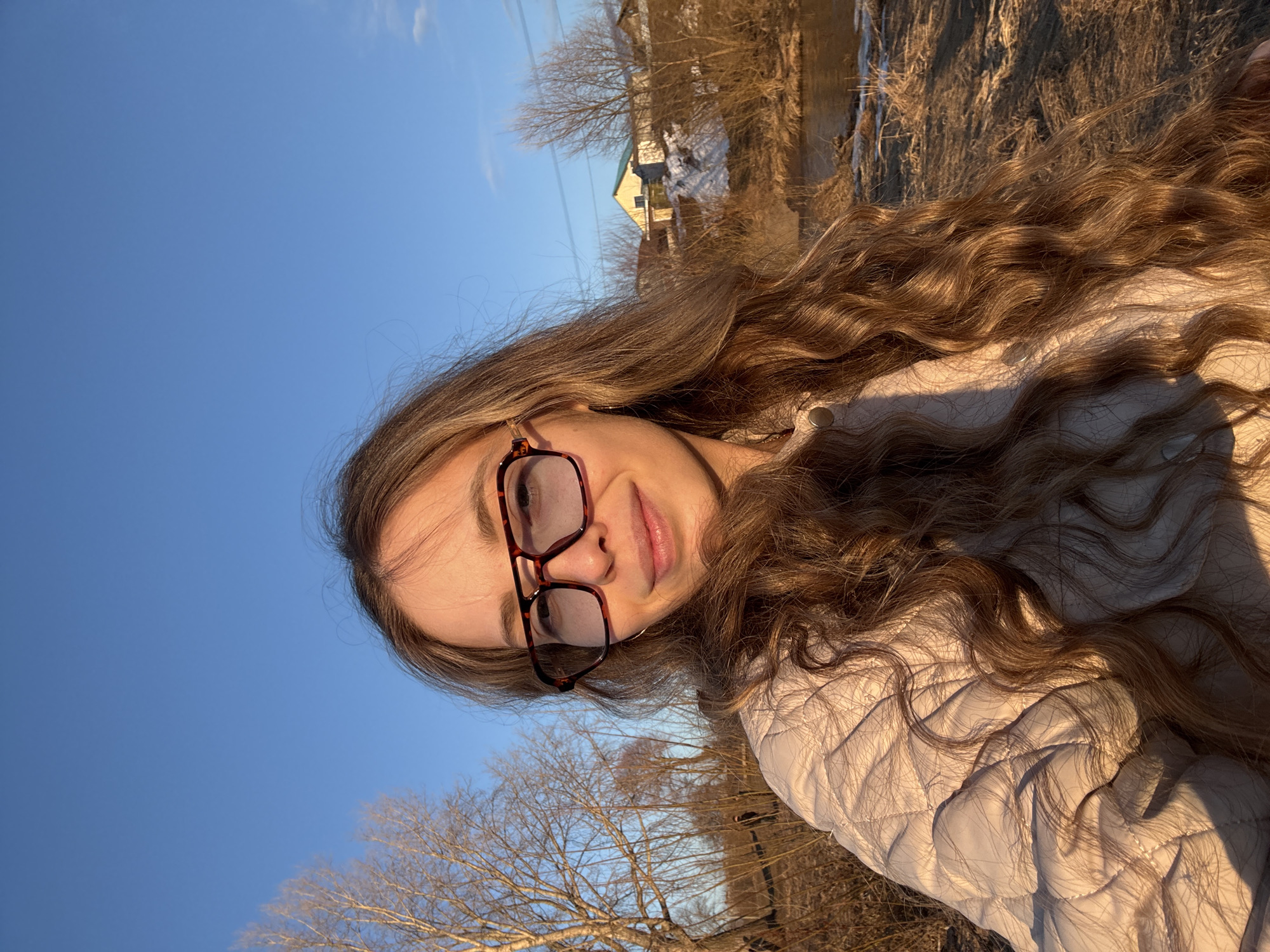 A young woman with long, wavy brown hair, wearing eyeglasses, is photographed outdoors in Chaikovsky, Russia. She wears a light-colored, quilted jacket. The background includes a clear blue sky, bare trees, a small stream or river, and a house partially visible. The photograph is taken at an angle, with the woman's face slightly tilted towards the camera. The scene appears to be illuminated by sunlight.