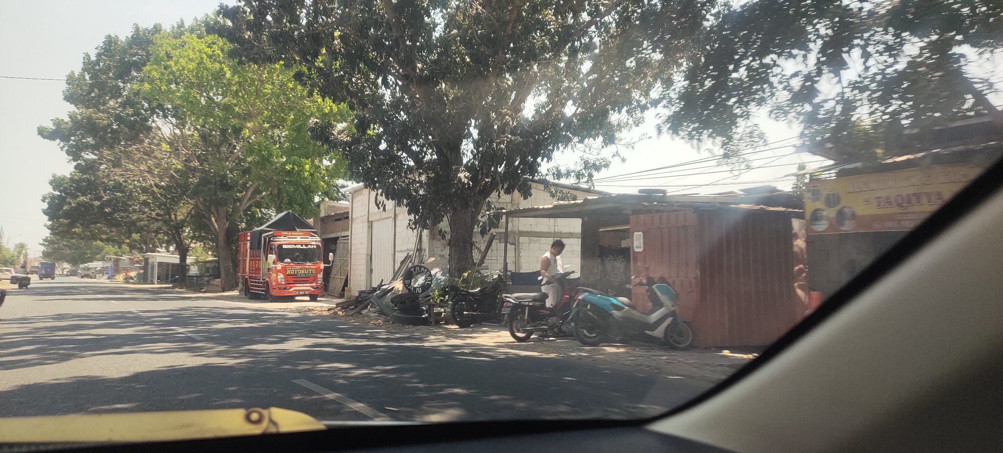 The image depicts a street scene in Mranak, Indonesia, on a sunny day. In the foreground, a yellow object, possibly part of a car's interior or dashboard, is visible. The street itself is paved and casts shadows from overhead trees. On the right side of the street, a red truck with a black canopy and white lettering that reads "BISMILLAH NOYOSUTO" is parked. Next to the truck, there is a cluster of what appears to be discarded items, including tires and debris. Further down the street, a covered structure with corrugated metal walls and an open front is situated. A person wearing a white shirt and pants stands near a motorcycle in front of this structure, interacting with it. Two scooters are also parked near this area. Beyond this, other buildings and trees line the street, with a yellow sign visible on one of the buildings that contains text including "TAQIYYA". The sky is bright and clear.