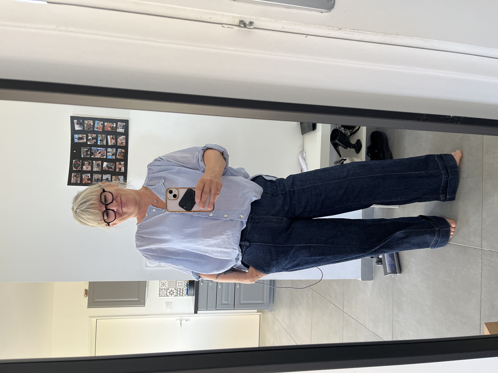 A woman stands in front of a mirror, taking a photo of herself with her phone. She has short blonde hair and wears glasses, a light blue striped shirt, and dark blue jeans. The reflection shows a portion of a room with white walls and gray cabinets. There's a collage of photos on the wall. The floor is tiled in a light gray color. A pair of black shoes sit on a shelf. The image was taken indoors. While Chambœuf, France is a possible location for where this image may have been taken, this cannot be confirmed from the image alone.