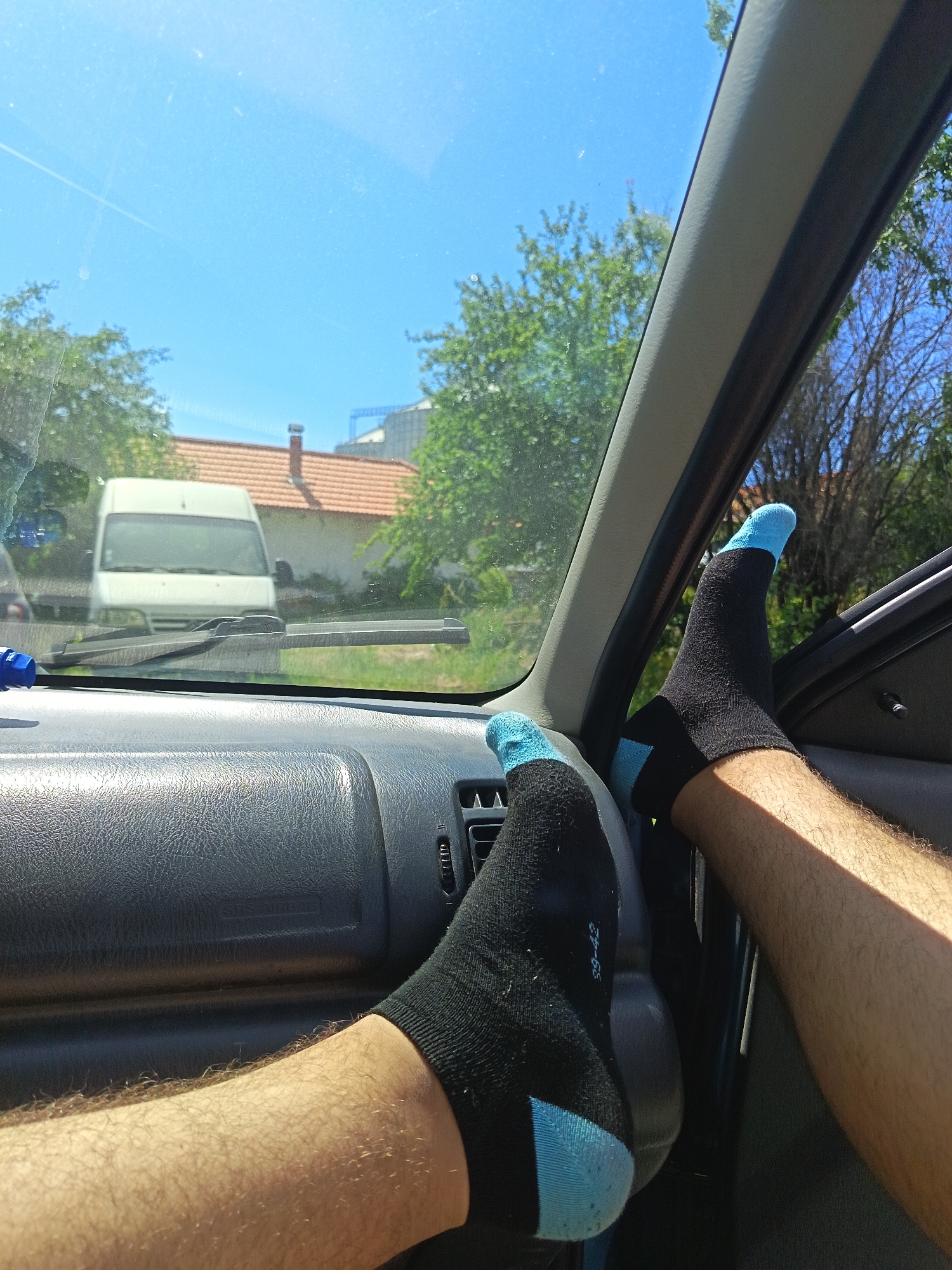 Inside a car, a person has their bare legs propped up on the dashboard, wearing black socks with blue toes and heels. Through the windshield, a white van is visible parked across the street from a house with a red tiled roof. Green trees line the background under a bright blue sky. The setting is Drenovets, Bulgaria.