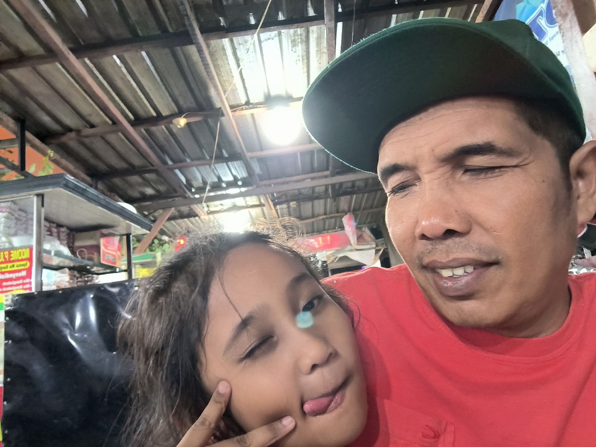 A close-up selfie shows a man and a young girl in what appears to be a casual indoor or semi-outdoor setting, possibly a market stall or small shop in Banda Aceh, Indonesia. The man, on the right, wears a dark green baseball cap and a red t-shirt. His eyes are closed or squinting with a subtle smile, conveying a relaxed or happy expression. The young girl, positioned on the left, has dark, wavy hair and is captured in a playful pose, winking with one eye, sticking out her tongue, and lightly touching her cheek with her fingers. A blue-green lens flare from a bright light source is visible on her face.
The background features a rustic ceiling constructed from wooden beams and planks, suggesting a simple, utilitarian structure. Several artificial light sources are visible, one of which is particularly bright and contributes to the lens flare. To the left of the girl, a black surface, possibly a display counter, is present. Further back, shelves and various items are partially visible, indicating a commercial environment. A red sign with mirrored, illegible white text, possibly "MOON" and "UL P", can be seen on the far left, along with a blue sign behind the man's head with unreadable text. The scene captures a moment of playful interaction and affection between the two individuals.