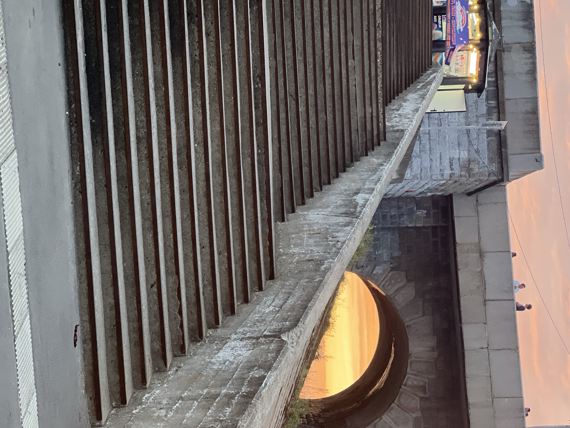 The image depicts a wide, concrete staircase with rusted metal edges, leading upwards. To the left, at the top of the stairs, is a small kiosk with a colorful awning displaying text in Cyrillic, appearing to sell food or drinks. The kiosk is illuminated, and a white door is ajar. Behind the kiosk, the stonework of a large structure is visible.
Above and behind the staircase, a concrete bridge spans across the frame. Several individuals are standing on the bridge, silhouetted against a vibrant orange and pink sunset sky. The bridge features a large arched opening, through which the sunset is prominently visible, casting a warm glow on the surrounding vegetation. The scene suggests an urban environment in Tver, Russia, likely a public space with a pedestrian overpass or bridge. The overall mood is serene, with the dramatic sunset providing a striking backdrop to the architectural elements and the daily life unfolding on the bridge.