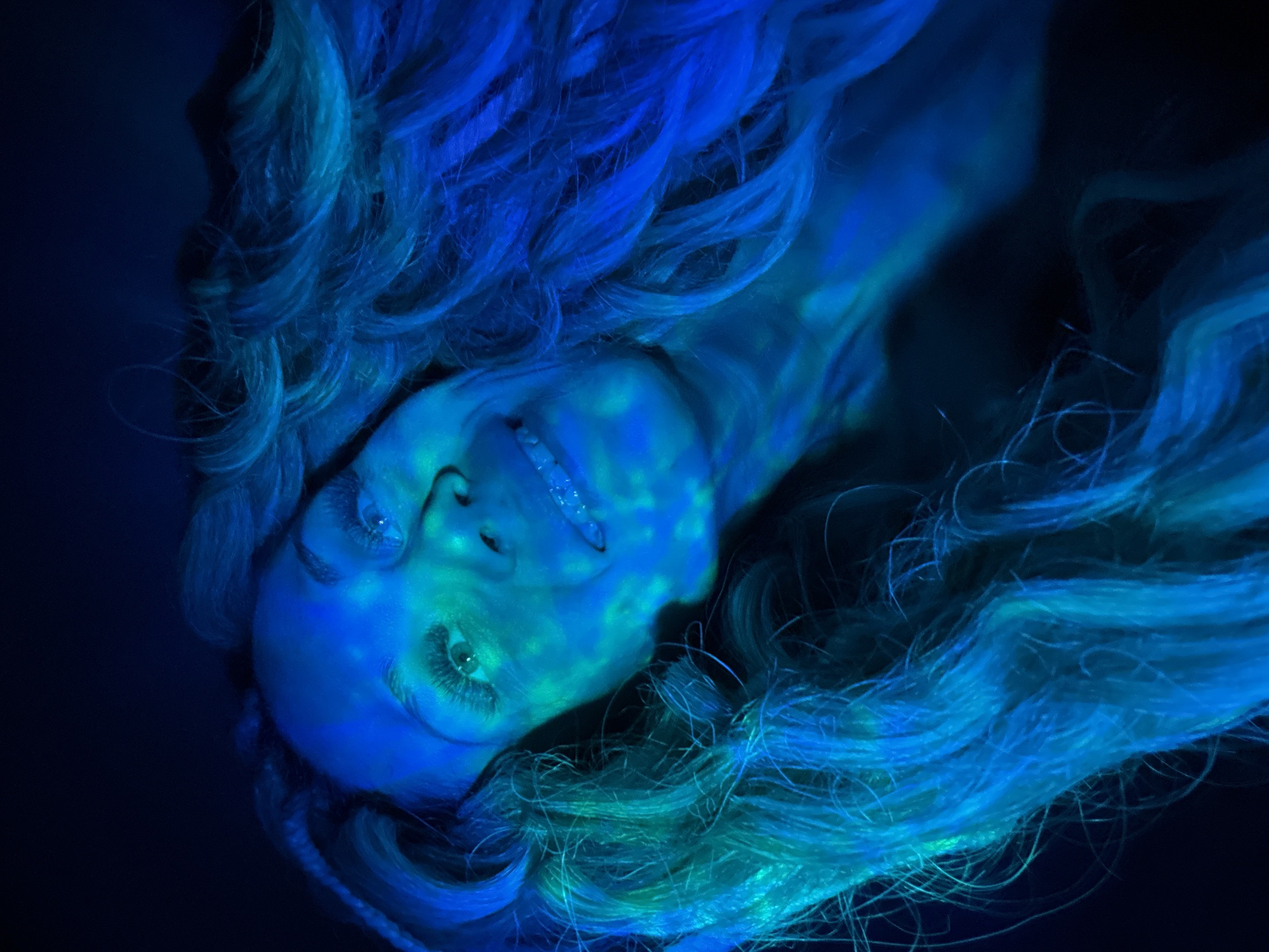 The image shows a person with light skin and long, wavy hair that appears blue and turquoise due to lighting. The person is looking up at the camera, smiling slightly, with their head tilted to the side. The face is illuminated with a bluish-green light, which creates shadows and highlights, emphasizing the facial features. The hair is spread out around the head, filling much of the frame. The background is dark, with little to no detail visible. No location information about Rusanovo, Russia is provided in the image.
