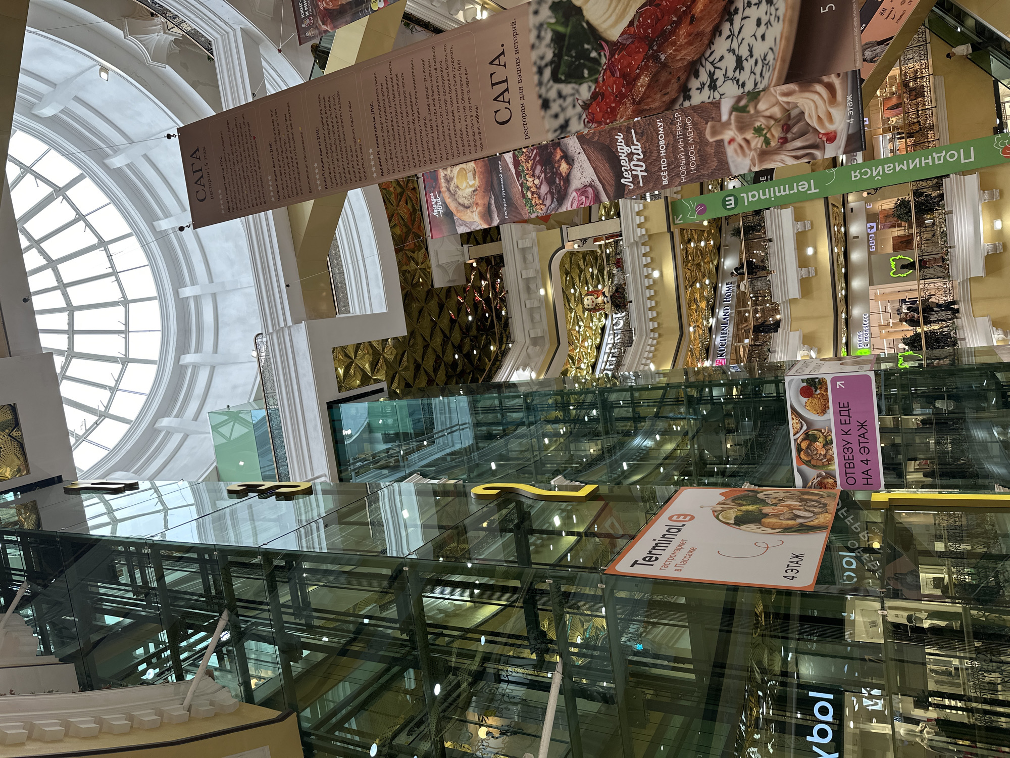 The image depicts the interior of a shopping center in Yekaterinburg, Russia. Banners advertising various restaurants are suspended from the ceiling. The name of one of the establishments, "SAGA," can be seen prominently, along with promotional material and menus. The shopping center features glass elevators.