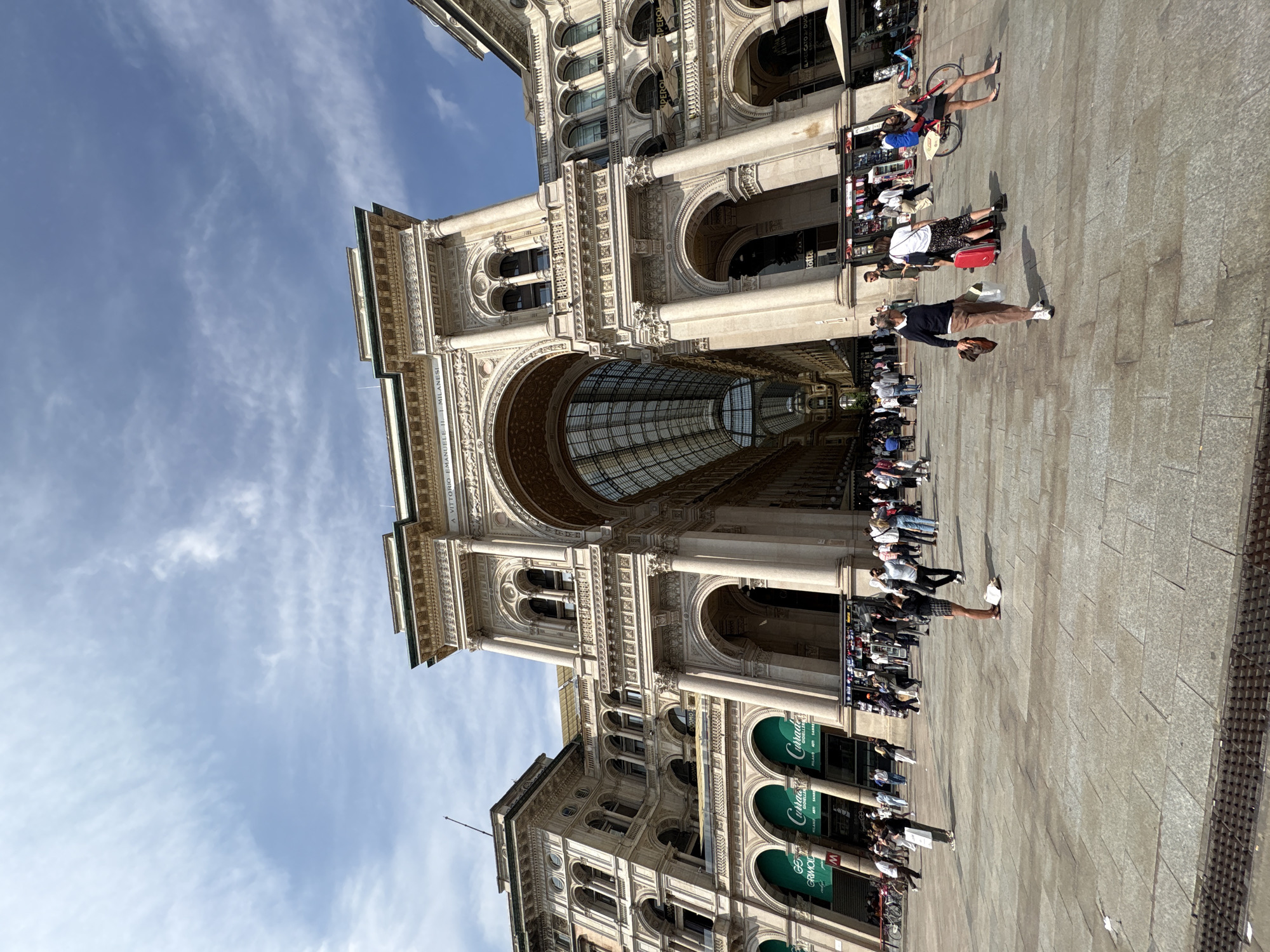 This image depicts the grand facade of the Galleria Vittorio Emanuele II in Milan, Italy, on a bright and sunny day. The monumental building, a prominent example of 19th-century architecture, dominates the frame with its ornate classical features.
The Galleria features multiple stories, characterized by large arched entrances, tall columns, intricate carvings, and balustraded windows. A distinctive glass and iron barrel-vaulted roof, covering the interior passage, is visible through one of the main arches. The stone facade appears to be made of light-colored material, likely marble or similar stone, with details highlighted by the direct sunlight.
In the foreground and midground, a significant number of people are visible on the paved plaza outside the Galleria. They are engaged in various activities typical of a bustling city center: walking, standing, and congregating. Some individuals are carrying shopping bags, indicating the area's commercial nature. Notable objects among the crowd include a red suitcase and several bicycles parked or being moved. The bright sunlight casts crisp shadows of the people and the architectural elements onto the paving stones, suggesting a time around midday or early afternoon. The sky above is a clear blue with scattered, wispy white clouds.
Visible text on the building's facade includes "GALLERIA VITTORIO EMANUELE II" and "MILANESE." Additionally, storefront signs with green awnings display text such as "CARRARO" and "GRIFO," indicating businesses located within or near the Galleria. The overall scene conveys a vibrant and lively atmosphere typical of a major European landmark and shopping destination.