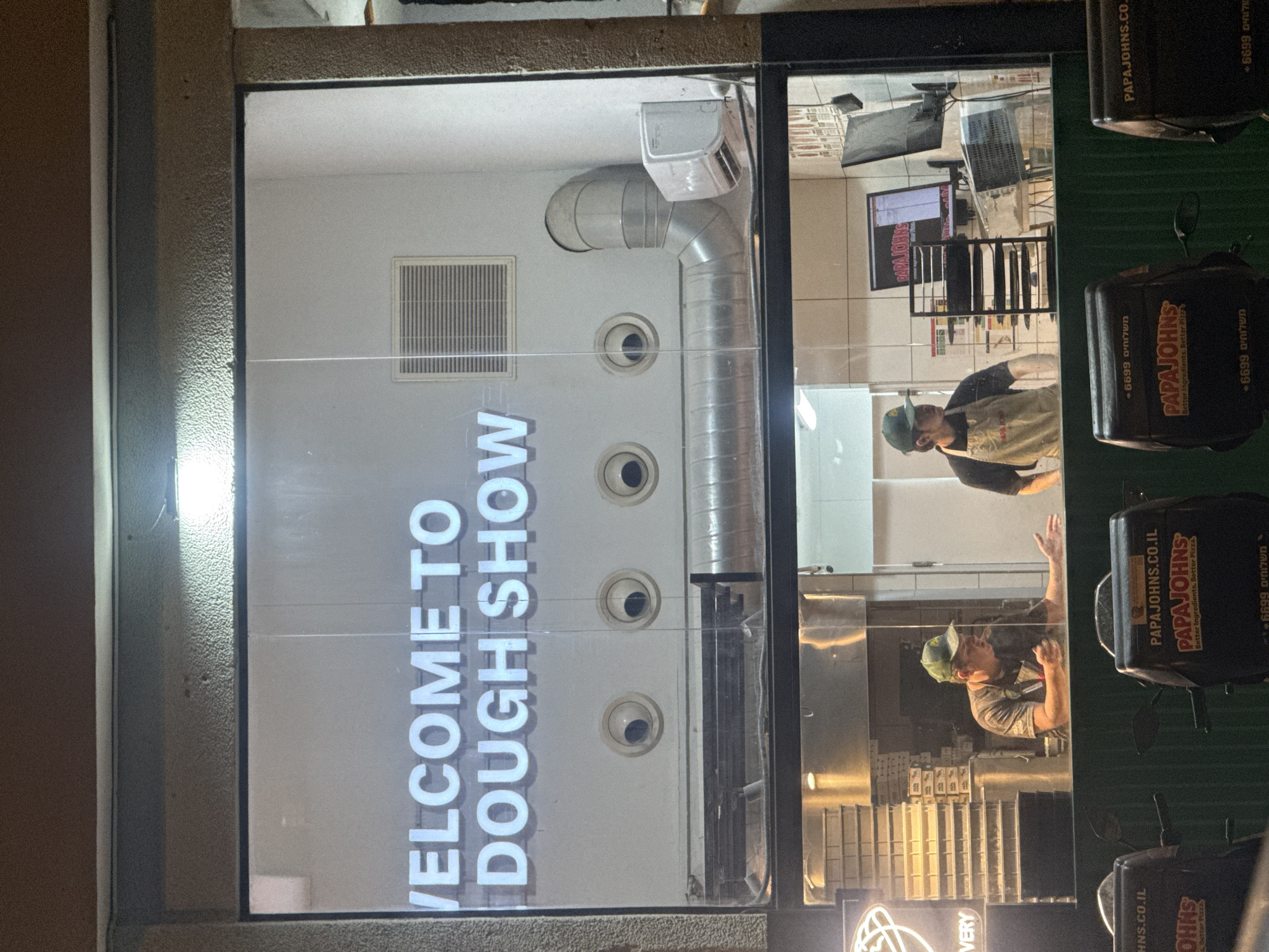 The image depicts the exterior of a Papa John's restaurant, likely a location in Ramat Gan, Israel, given the Hebrew script on the delivery boxes. The scene is viewed through a large window displaying the words "WELCOME TO DOUGH SHOW." Inside, two employees, both wearing hats and aprons, appear to be working or interacting behind the counter. A ventilation system with a large metal duct runs along the wall, punctuated by circular vents. The restaurant's interior includes shelves, a computer monitor, and other equipment typical of a food service establishment. Delivery boxes branded with the Papa John's logo and phone number are affixed to parked delivery scooters or motorcycles outside, suggesting active delivery operations.