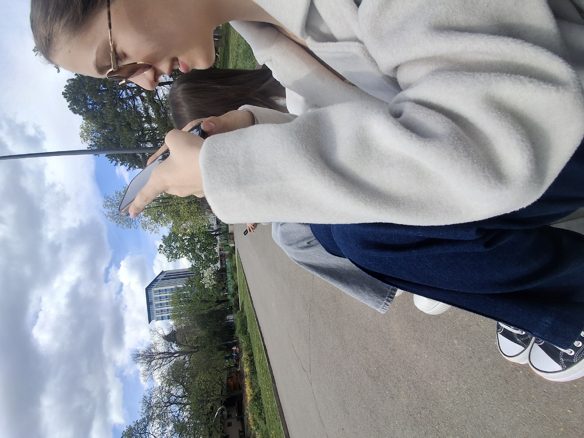 A young woman with sunglasses is sitting outside in a park. She is wearing a white hoodie and dark blue pants, with black and white sneakers visible. She is looking at and using a smartphone. Another person with dark hair is sitting next to her, partially visible. The location appears to be a public park in Sofia, Bulgaria, based on the presence of a modern high-rise building in the background, park landscaping, and trees. The sky is partly cloudy.