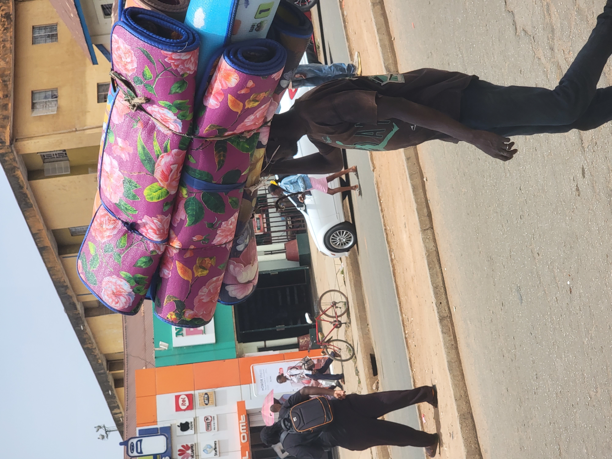 The image captures a bustling street scene in Jos, Nigeria, on a clear, sunny day. The central figure is a dark-skinned man walking on the paved road, expertly balancing a large stack of rolled-up mats on his head and shoulder. The mats are vibrantly colored, primarily purple with a distinct pink floral pattern resembling roses, alongside some blue mats. He wears a dark t-shirt with a green design and dark trousers, with a ring visible on his left hand.
In the background, several buildings line the street. One prominent building is yellow-brown with multiple windows. Further down, another building features distinct orange and green sections, displaying various commercial logos such as "Itel," "Apple," "Huawei," "Samsung," "MTN," "StarTimes," and "Free TV," suggesting a mobile phone or electronics store.
Vehicles are present, including a white car parked on the side of the road and a red bicycle propped up nearby. Other individuals are visible in the scene: a person in a light top and pink skirt near the white car, another person holding what appears to be an umbrella further down the street, and a person in dark clothing with a black backpack and orange trim walking away in the foreground. The asphalt road and concrete curb complete the urban setting, illustrating typical street commerce and daily life.