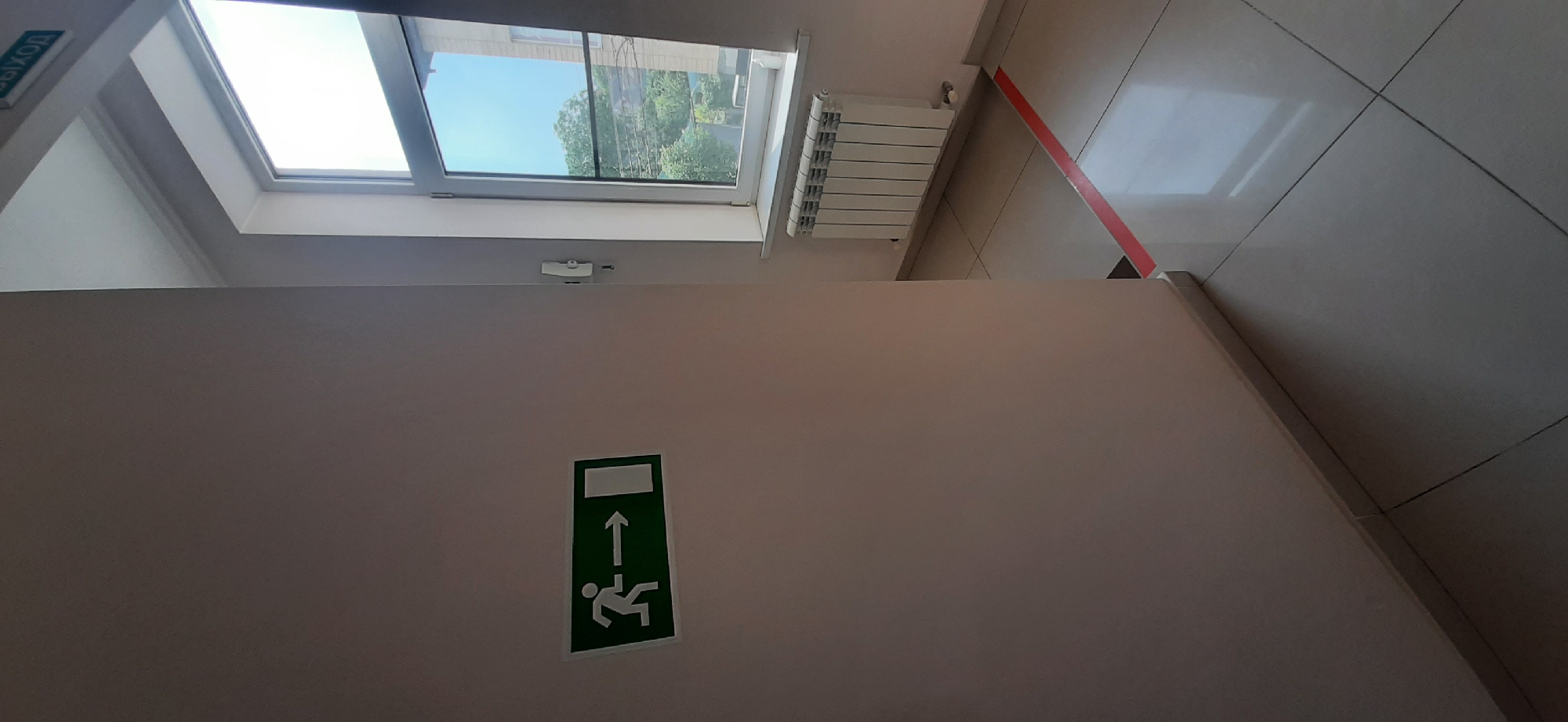This media file captures an interior view of a building, likely a stairwell or hallway, in Yessentuki, Russia. The scene is dominated by a large, light-colored wall that occupies the majority of the frame.
Mounted on the wall is a green and white pictogram sign. The sign depicts a figure in motion, with an arrow pointing upwards, suggesting an emergency exit or evacuation route. Above the sign, a white rectangular object with a lever is visible, possibly a door closer or a latch mechanism.
In the upper portion of the image, a skylight or large window allows natural light to flood the space. Through the window, trees and a blue sky are visible, indicating it is daytime. Adjacent to the window, a white radiator is mounted on the wall.
The floor appears to be tiled in a neutral color, with a red stripe running diagonally across it in the upper right portion of the image, potentially marking a walkway or safety zone. At the top left of the frame, a sign with Cyrillic text is partially visible, reading "ВЫХОД," which translates to "EXIT" in Russian. The overall impression is one of a functional, perhaps utilitarian, indoor space.