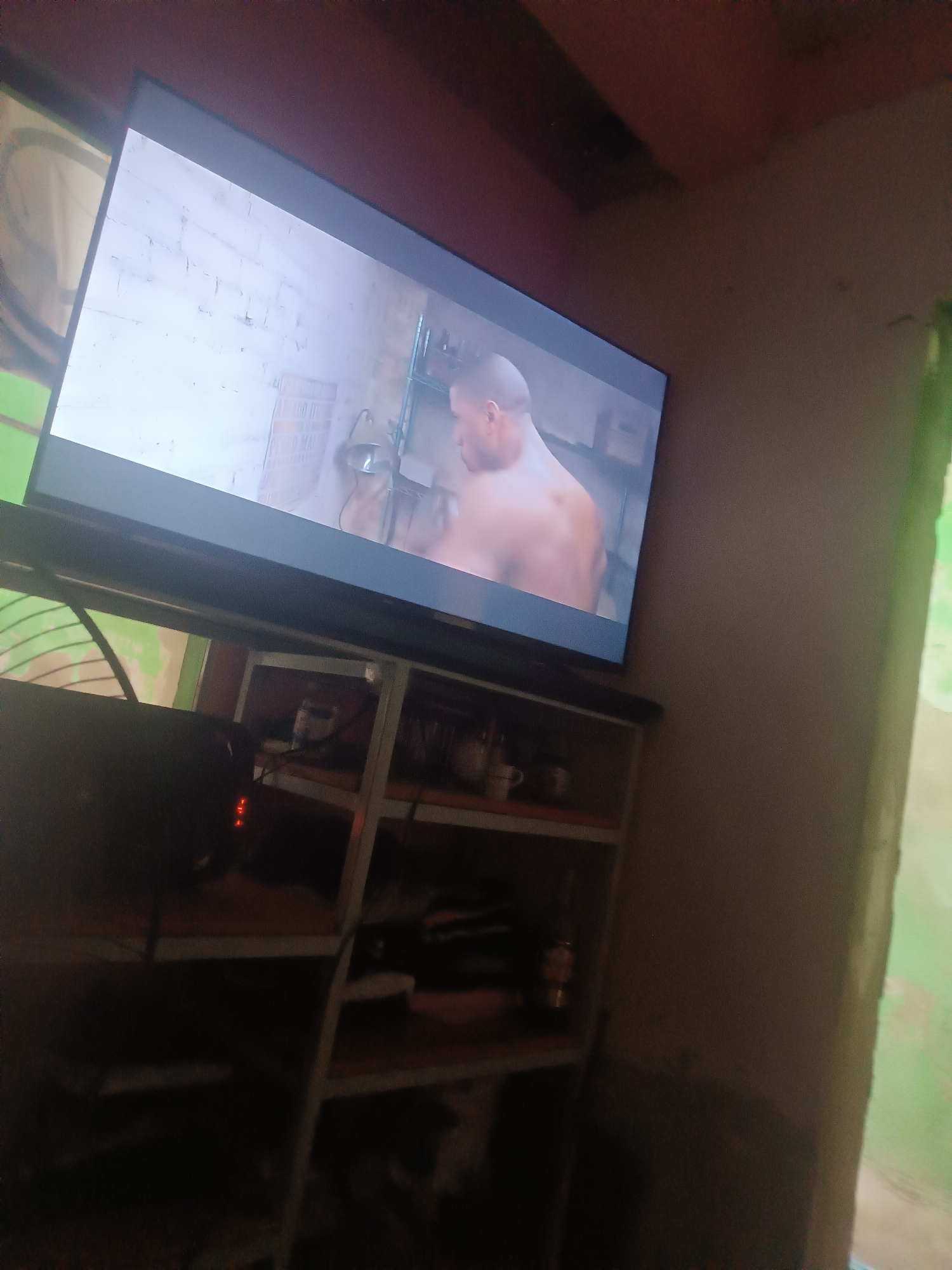 A television displays a scene of a shirtless man with shaved head, possibly in a bathroom setting with a white brick wall, metal shelving, and a desk lamp. The television is placed on a metal shelf with various items on its shelves including cups and what appear to be clothes. The location is indoors and appears to be in Porlamar, Venezuela.