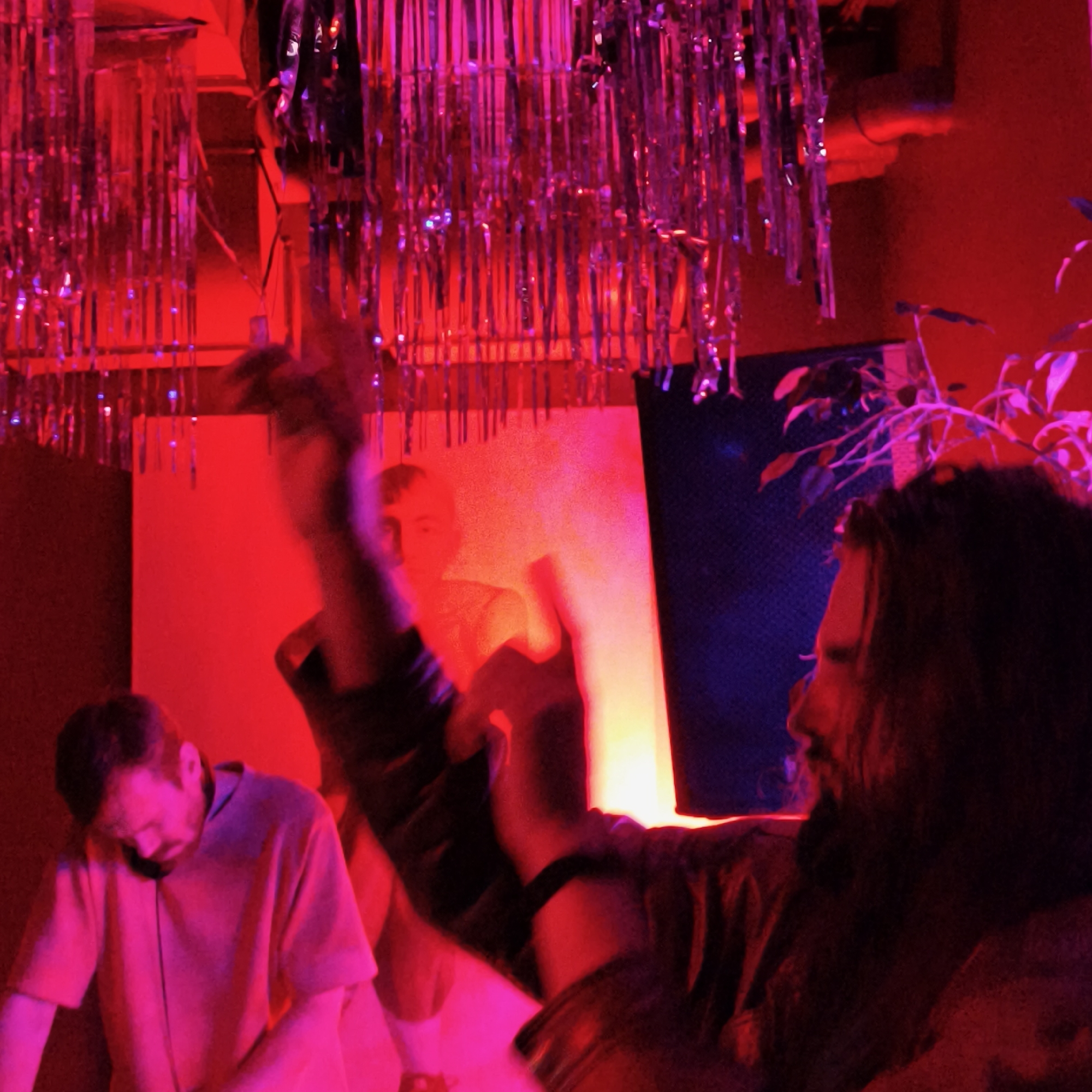 The scene, likely within a nightclub or entertainment venue in Hrodna, Belarus, is bathed in intense red and pink lighting, punctuated by strands of metallic fringe hanging from the ceiling. In the foreground, a person with dark, long hair and a beard is seen in profile, raising their hands. A wristwatch is visible on their wrist. To the left and in the background, a second person, likely a DJ, is seen wearing headphones and tending to equipment. A square illuminated surface is behind them. Foliage from a plant is visible near the right edge of the frame.