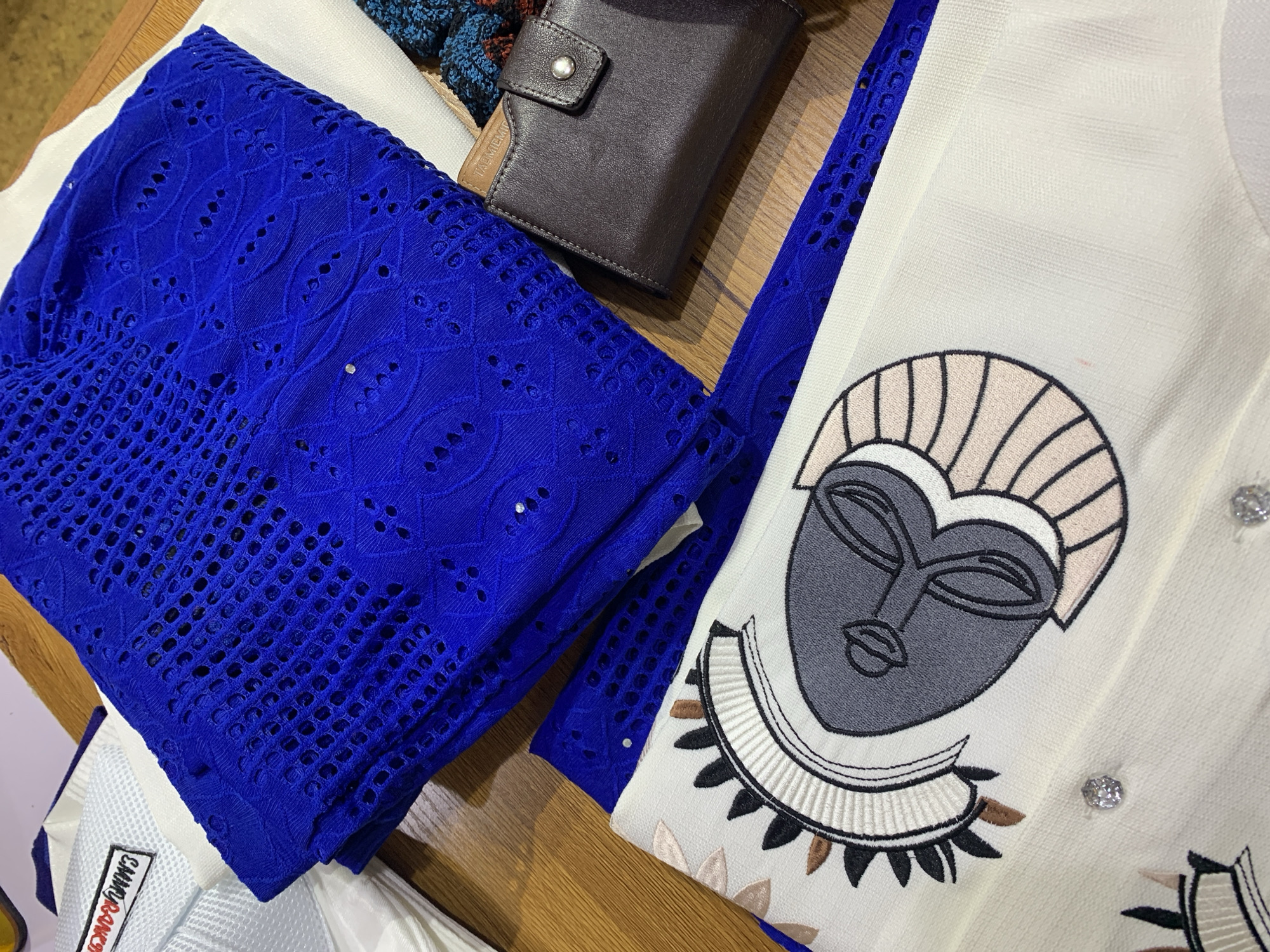 The image shows a close-up, overhead view of fabric and accessories on a wooden surface, likely a table, in a setting that could be within Gura Top, Nigeria. In the center and top of the frame, a stack of bright blue, intricately patterned lace fabric is prominently displayed. To the left of the blue lace, a white mesh hat with a black and red embroidered tag reading "COMMERCIAL" is visible. Below the blue lace and extending towards the bottom of the frame is a piece of cream-colored fabric with a detailed, embroidered design of a stylized mask or face, featuring black, gray, white, and brown elements. Scattered on this cream fabric are small, reflective embellishments, likely rhinestones. To the right of the blue lace and slightly behind it is a dark brown leather wallet with a silver snap closure. The wooden surface appears to be a table with a visible grain. No people or actions are depicted in the image.