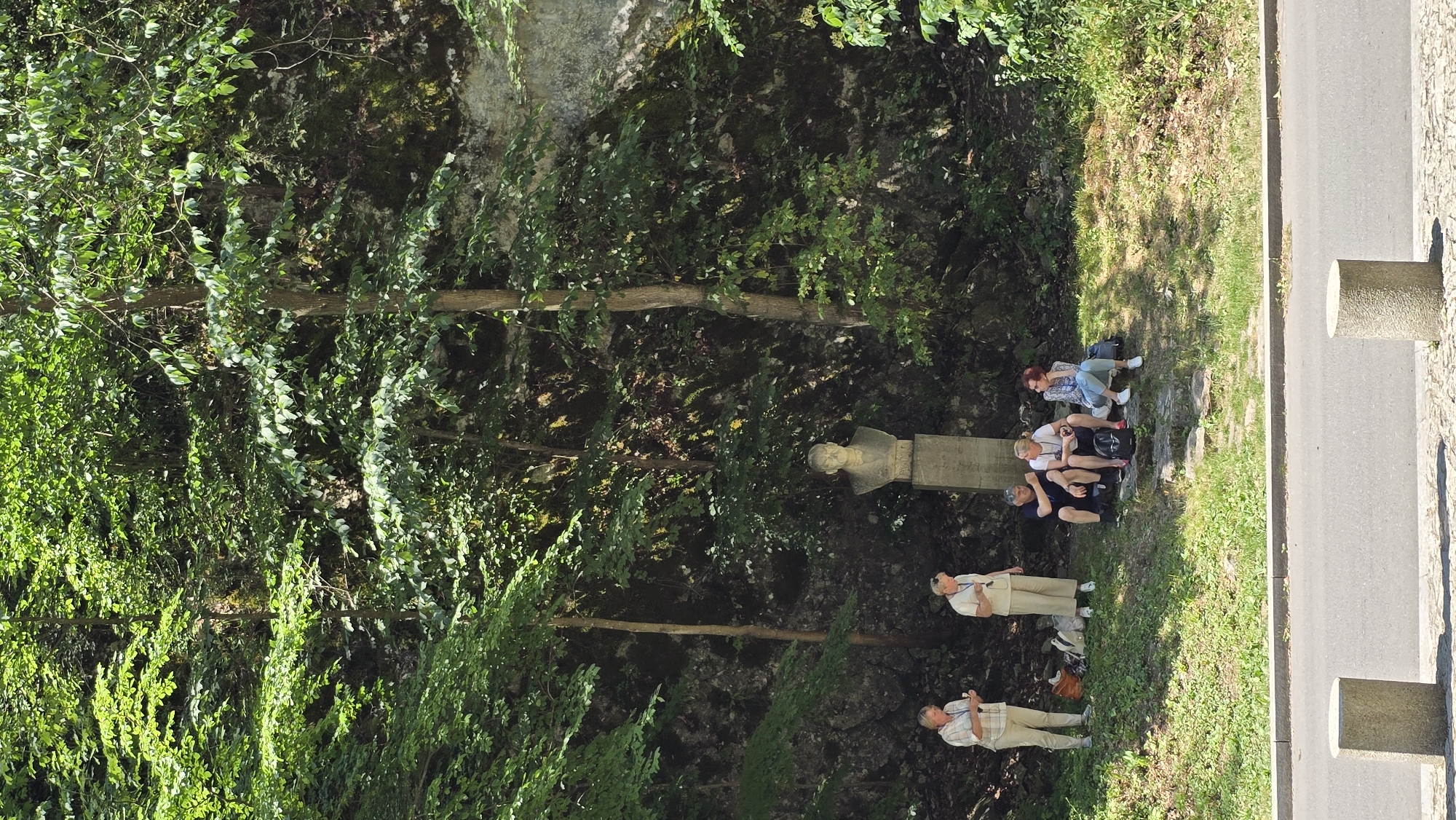 The image depicts a group of people resting outdoors in a lush, green environment, likely a park or natural area, near a road. A stone bust or statue is visible, suggesting a historical or memorial site. The people are dressed casually, indicating a relaxed atmosphere. Some are sitting on a stone ledge, while others are standing. One person is holding a camera, possibly taking photos. The scene appears to be taking place during the daytime, with bright sunlight casting shadows. The presence of trees, foliage, and a rocky incline suggests a natural setting. The location is identified as Vavřinec, Czechia.