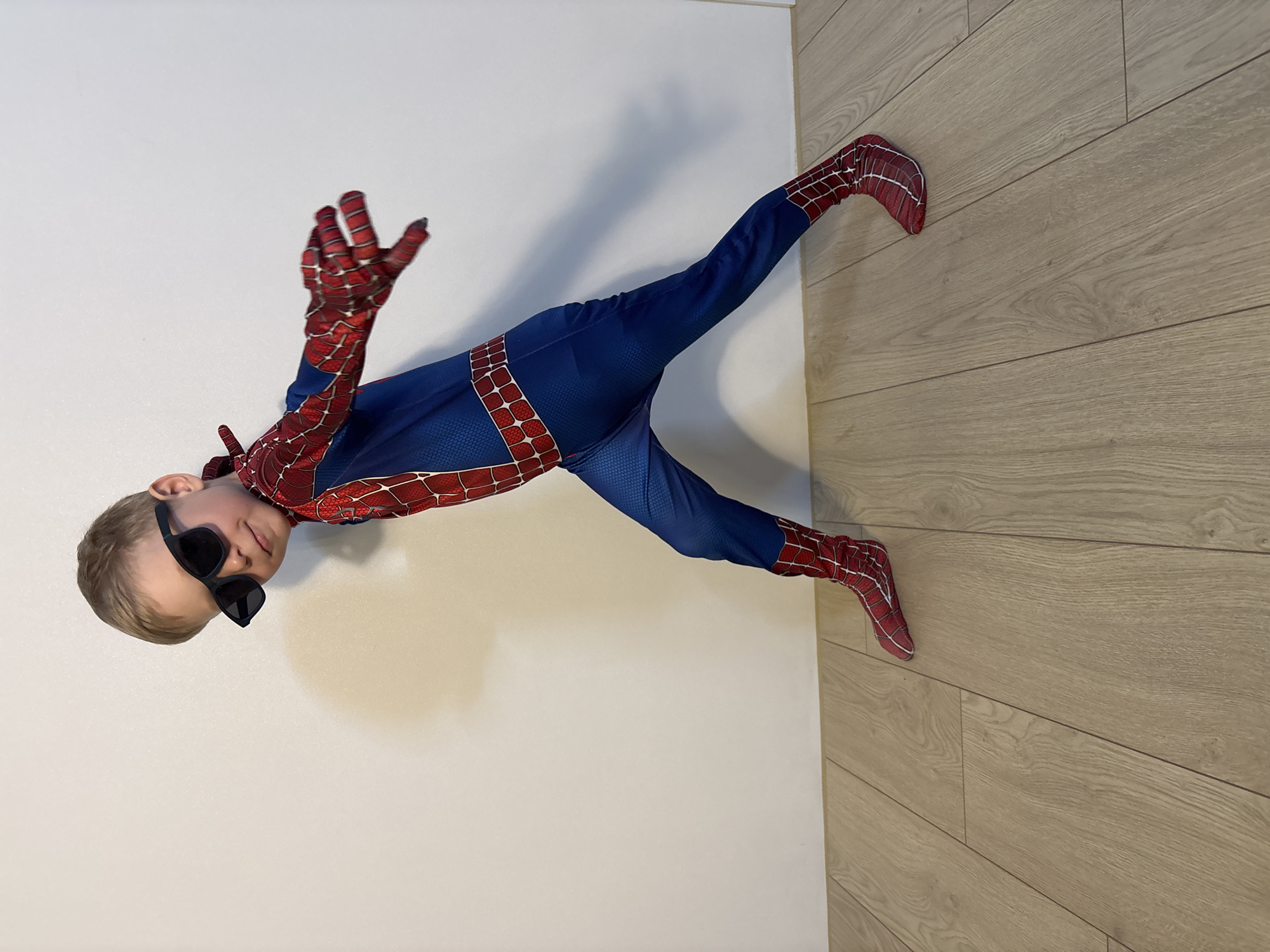 A young child, dressed in a Spider-Man costume, poses indoors. The child is wearing black sunglasses and is positioned diagonally in the frame, with the head near the left edge of the image and the feet extending towards the lower right. The background is a plain white wall on the left and a wooden floor on the right. The location is Chaikovsky, Russia, based on prior information.