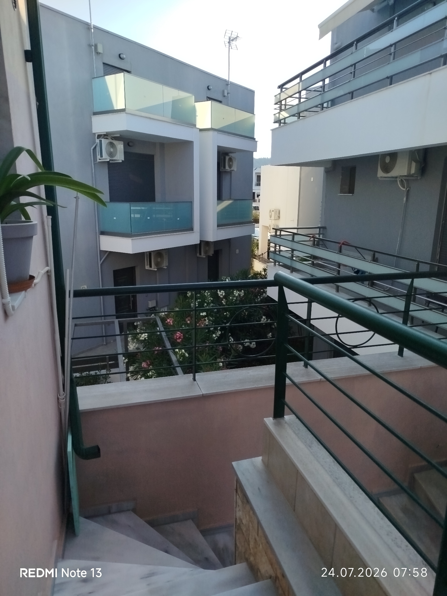 The image shows an outdoor view of a residential building complex, likely in Sithonia Municipal Unit, Greece, based on the timestamp. The perspective is from a staircase looking up towards balconies and upper levels of two adjacent buildings. The building on the left is painted light gray and features modern balconies with glass railings. An air conditioning unit is visible on the exterior wall. The building on the right, also gray, has similar balconies and a walkway with a ramp. Trees with flowering bushes are visible between the buildings. The foreground shows a set of marble stairs leading upwards, with a pinkish-colored wall and a dark green drainpipe on the left. The sky is clear and light blue. The timestamp "24.07.2026 07:58" and "REDMI Note 13" are present in the lower corners of the image. There are no people or animals visible in the scene.