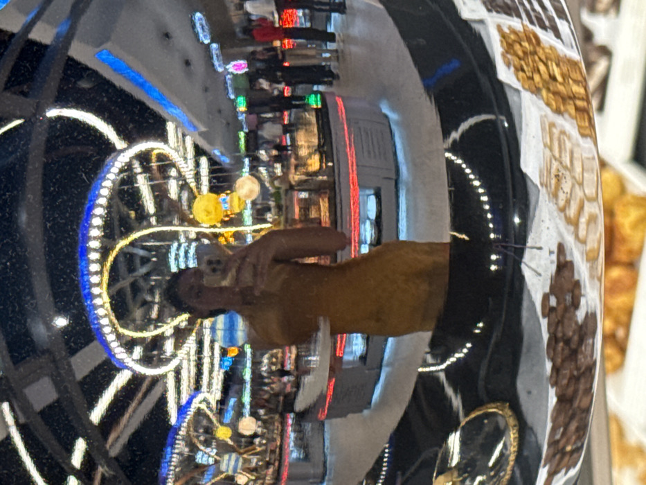 The image shows a distorted reflection in a black, possibly metallic surface. The reflection reveals a person in a yellow shirt taking a picture with their phone. Behind the person, illuminated lights decorate the surrounding area. Various food items appear on the right side.
