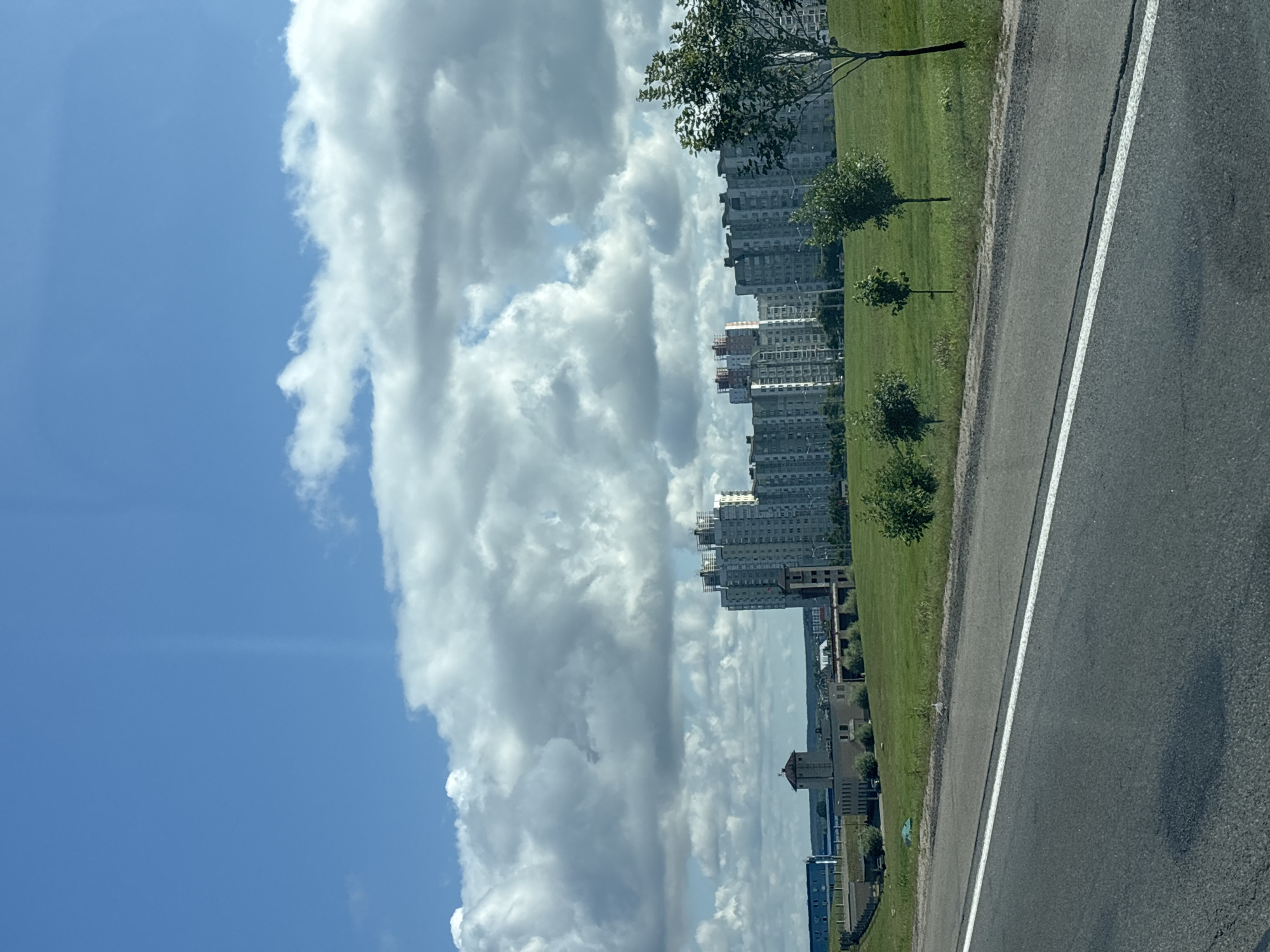 The image captures a view of a road leading towards a suburban landscape under a bright, cloudy sky. In the mid-ground, a row of multi-story apartment buildings stretches across the horizon, characterized by their uniform, grey facades and numerous windows. To the left of these buildings, a distinct, square tower with a pointed roof stands out, possibly a remnant of older architecture or a functional structure. Further left, a long, blue building with a modern design is visible, suggesting an industrial or institutional presence. The foreground is dominated by a paved road with a white line marking its center, and to the right, a green, grassy field with scattered trees. In the distance, the landscape appears flat, with more buildings and trees forming the backdrop. A single bird is present in the field, near the edge of the road. The overall scene suggests a developing urban or peri-urban area. Based on the architectural style and the presence of numerous Soviet-era style apartment blocks, this location could be in a residential district of Minsk, Belarus, a city known for such urban development.