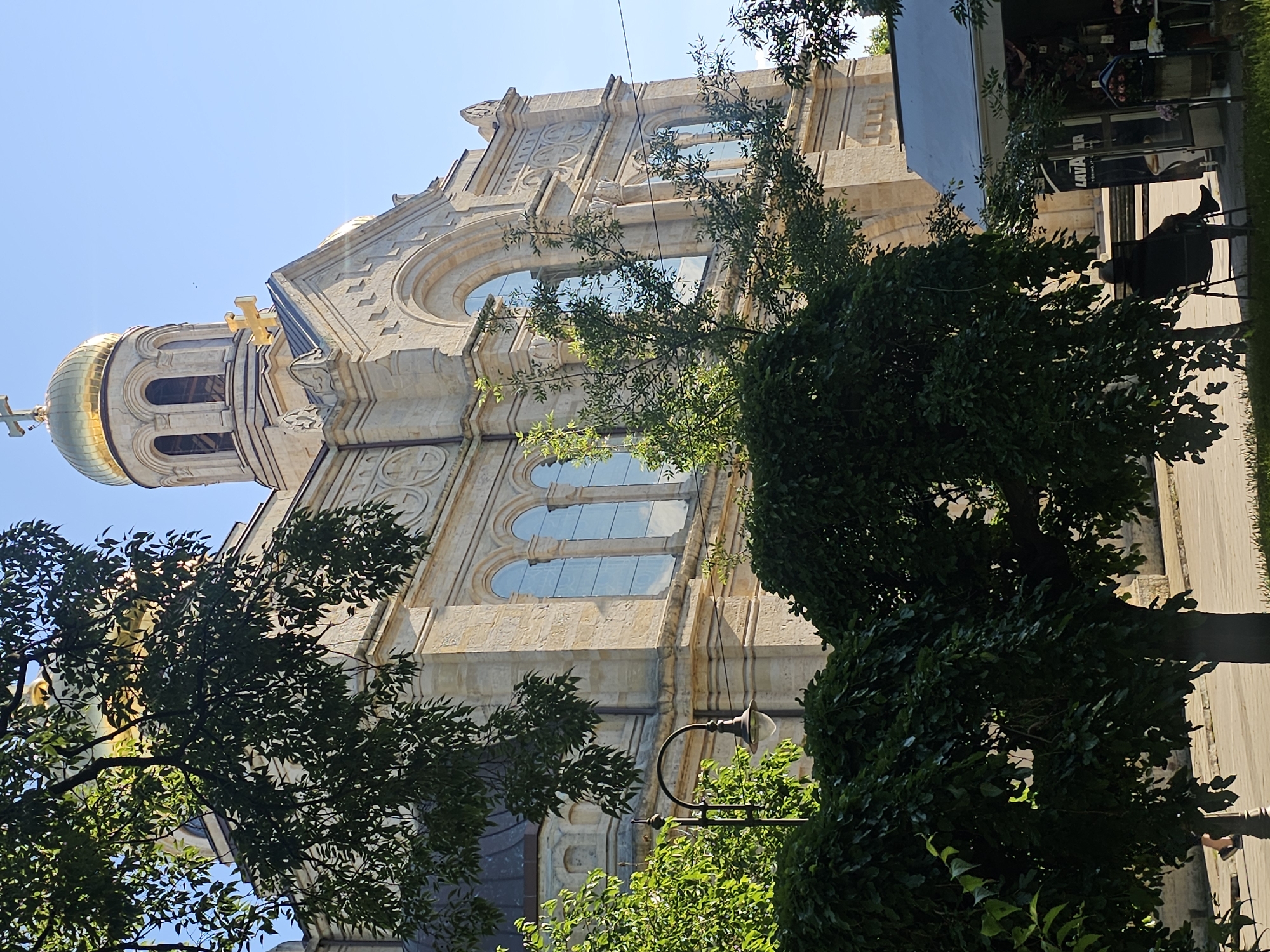 The image shows the exterior of a large, ornate stone building, likely a church, with golden domes. The building is made of light-colored stone and features intricate carvings and arched windows. Lush green trees and foliage partially obscure the lower portions of the building, with branches and leaves in the foreground. A street lamp with an ornamental design is visible among the trees. In the background, a small kiosk or shop with signage that includes the word "LAVAZZA" is present. A person is seated in a chair outside this kiosk. The scene is illuminated by bright sunlight, indicating daytime. The sky is clear and blue.