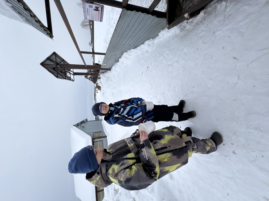 Two young males are depicted outdoors in a snow-covered environment identified as Zaosinovo, Russia, under an overcast sky. The ground is uniformly covered in packed snow, forming a clear path in the foreground.
The subject on the left, a pre-teen male, wears a camouflage-patterned winter suit (yellow, green, black, grey) and a dark blue knitted beanie with red star insignias. He holds a white plastic bucket in his right hand, looking downwards.
The subject on the right, a younger pre-teen male, wears a blue, black, and white patterned winter jacket, black winter pants with white reflective accents, a black knitted beanie, and glasses. He holds two white plastic buckets, one in each hand, and is looking towards the viewer.
To their right, a metal fence extends into the distance, featuring dark posts, a horizontal rail, and a lower section of corrugated metal sheeting. Overhead, several angled structures with corrugated metal roofs are visible, extending along the fence line. A white sign attached to a fence post displays text in Russian, including "Як" and "Бизон" (Yak and Bison), a QR code, and illustrations of these animals. A wooden feeding trough is positioned at the lower right, adjacent to the fence.
Beyond the fence is a snow-covered field, within which dark, indistinct shapes suggest the presence of large animals further down the enclosure. In the background to the left, two single-story buildings with snow-covered gable roofs and light-colored walls are visible. The distant horizon reveals a snow-covered landscape beneath a uniformly grey, overcast sky.
