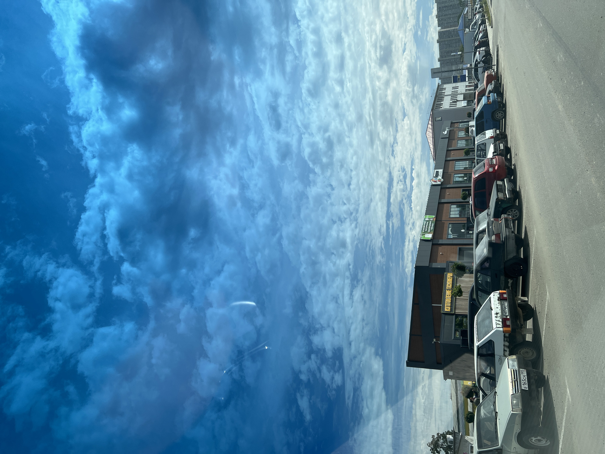 The image captures a bright, partly cloudy daytime scene in an urban setting identified as Krasnodar, Russia. The sky dominates the left side of the frame, showcasing a vast expanse of blue with dynamic formations of white and grey clouds, suggesting a mix of clear patches and more dense cloud cover.
The right side of the image features a street scene. A row of various vehicles, including sedans, SUVs, and vans, are parked along an asphalt road or parking lot. Several Russian license plates are visible on the cars, notably "Е165СН 93" and "В071НК 93", with the "93" region code confirming the location in Krasnodar Krai.
Behind the parked cars stands a multi-story commercial building with a modern design, characterized by dark grey and brown exterior elements. Several signs are affixed to its facade. On a green sign partially visible on the far left, Russian text reads "ПРОДУКТЫ" (Groceries), "СВЕЖАЯ" (Fresh), "ПТИЦА" (Poultry), and "ПОСУДА" (Dishes/Tableware). Another yellow sign on the building displays the text "Уютный дом" (Cozy Home). Further in the background, taller residential buildings indicate a developed urban environment. The scene appears static, depicting a typical day with no specific event or activity taking place. The lighting suggests mid-day conditions.