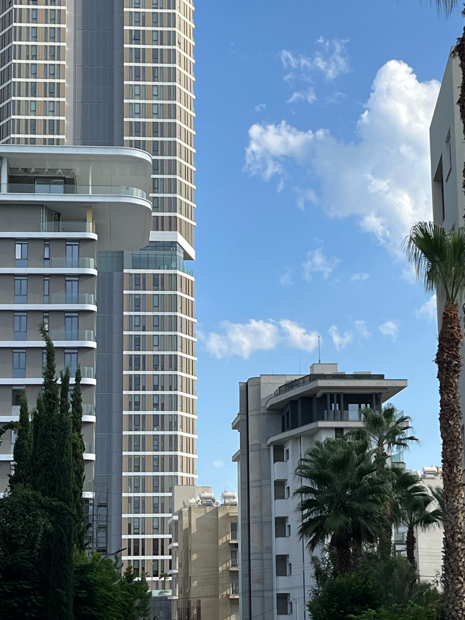 The image shows a street view in Limassol, Cyprus, with several modern buildings dominating the scene. In the foreground, tall evergreen trees line the lower portion of the frame. Behind them, a multi-story building with a light-colored facade and numerous windows is visible. To the right of this building, a palm tree with a textured trunk and green fronds is prominent.
The central focus is a very tall, slender skyscraper with a facade featuring alternating vertical sections of light-colored panels and windows. A distinctive curved balcony or terrace juts out from the midsection of this skyscraper. Adjacent to it, and slightly shorter, is another modern building with a more angular design and a dark-colored roof structure. This building also features multiple balconies and numerous windows. Further to the right, the edge of another building, partially obscured by the palm tree, is visible. The sky above is a bright blue with scattered white clouds, suggesting fair weather. No people are visible in the image. The overall impression is an urban environment with contemporary architecture.
