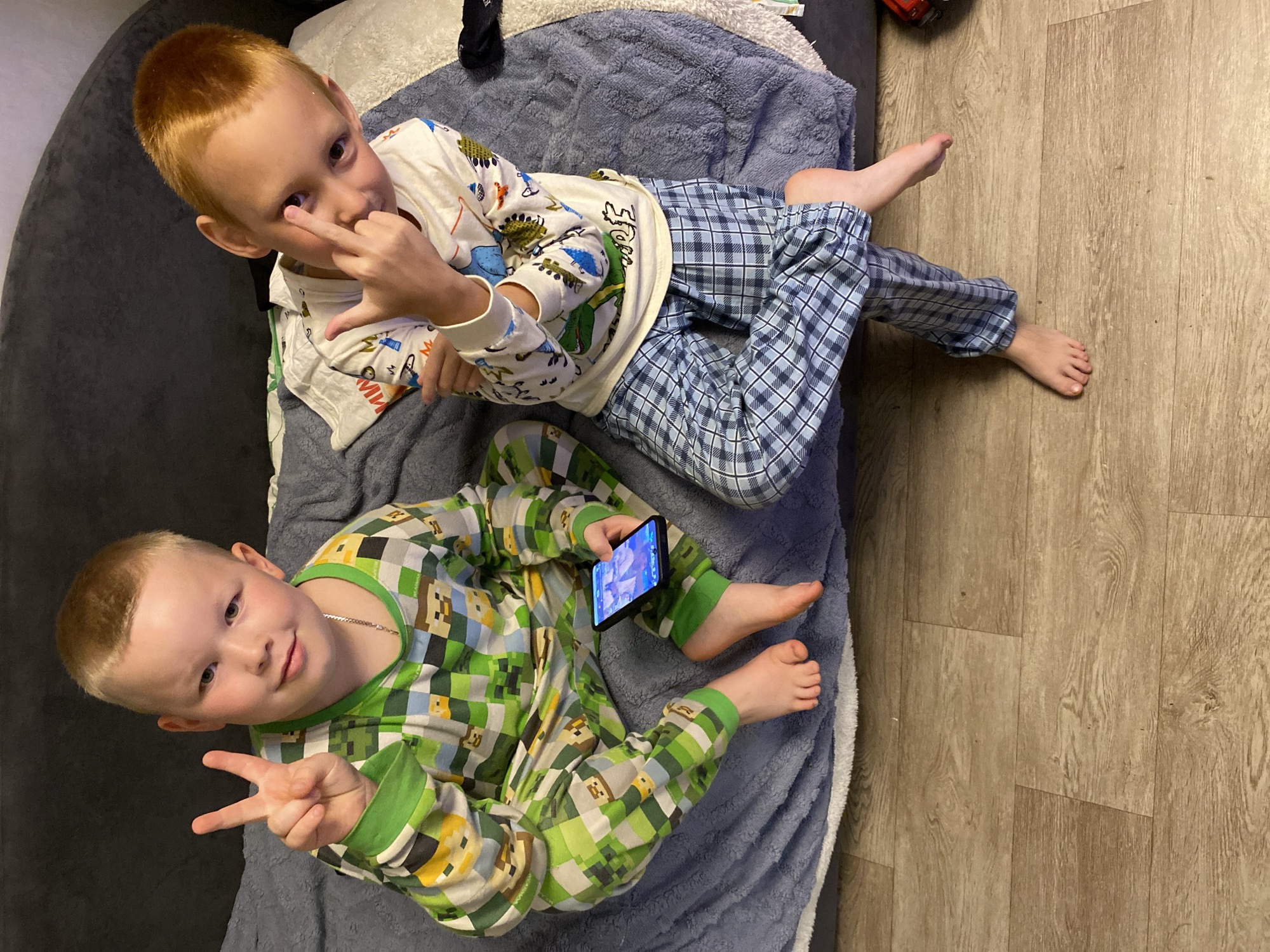 The image shows two young boys sitting on a bed or sofa covered with a grey blanket. The boy on the left, with light brown hair, is wearing green and white plaid pajamas with a Minecraft-themed pattern. He is holding a black smartphone in his right hand and making a peace sign with his left hand. The boy on the right, with reddish-blonde hair, is wearing white pajamas with dinosaur and other cartoon prints, and blue plaid pajama pants. He is looking at the camera and making a rude gesture with his right hand. They are both barefoot, and the wooden floor is visible beneath them. The scene appears to be indoors, likely in a home setting. There is no identifiable information in the image to suggest a specific location in Noviy, Russia.