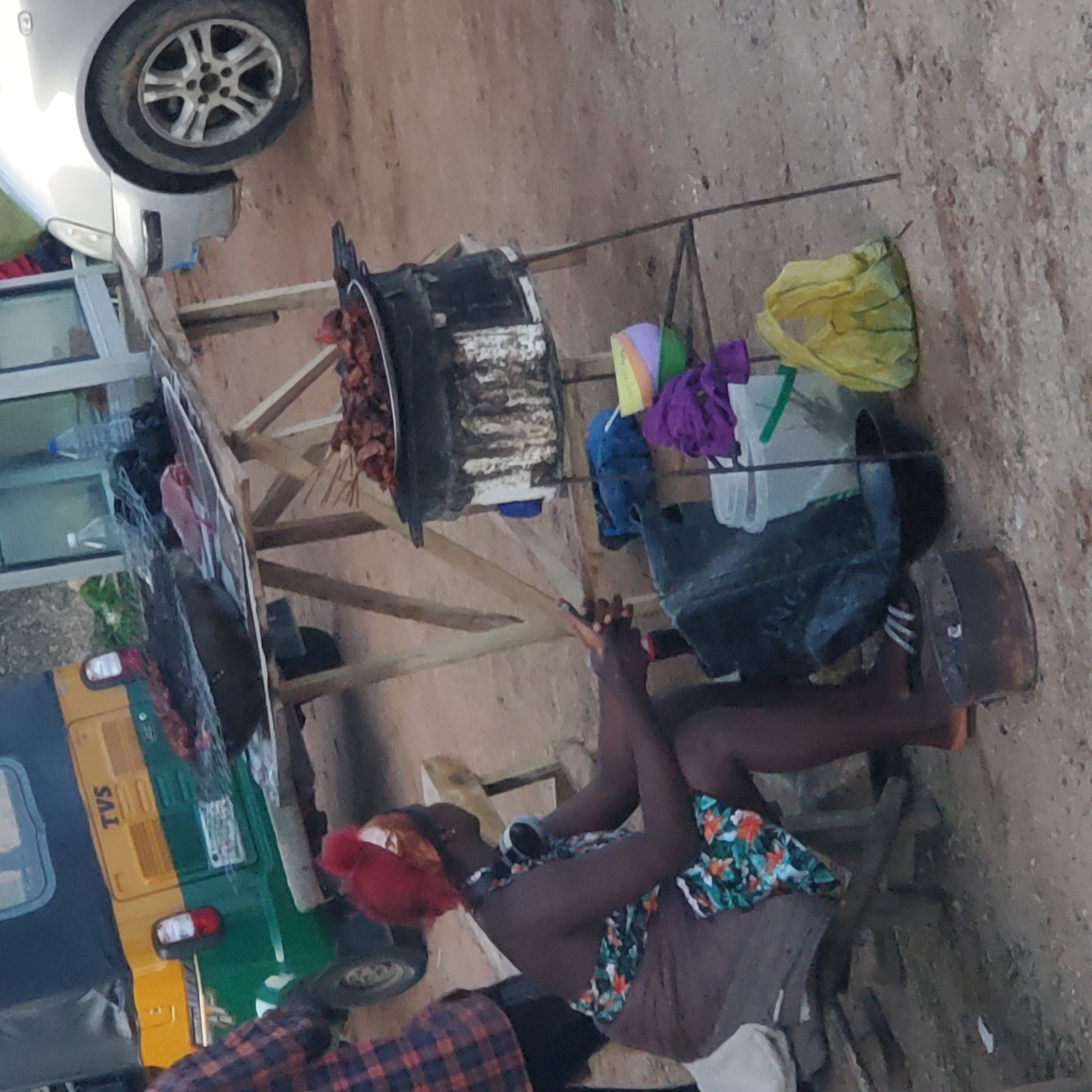 The image depicts an outdoor scene, likely on a sandy or dirt lot in Jos, Nigeria, where street food is being prepared and sold. In the foreground, a woman with dark skin, wearing a red headscarf and a floral-patterned wrap skirt, is seated on a low stool. She has headphones around her neck and appears to be in the process of preparing food or interacting with it. To her left, a rusty metal container sits on the ground, possibly a makeshift stove or brazier. Behind her, a black plastic container is visible.
A wooden table, precariously constructed from planks and branches, is set up in the middle ground. On this table, a metal grilling apparatus holds skewers of cooked meat. There are also some unskewed pieces of meat and a pinkish raw meat slice on a newspaper. Various items, including plastic bottles and black plastic bags, are present on and around the table. A portion of a three-wheeled auto-rickshaw, painted yellow and green with "TVS" branding, is visible in the background, suggesting it may be used for transport or as part of the stall. A silver car is parked to the right of the scene. The ground is a mixture of sand and dirt. A yellow plastic bag and other colorful containers are also scattered around the stall. The overall impression is of a busy, informal street-side food vending operation.