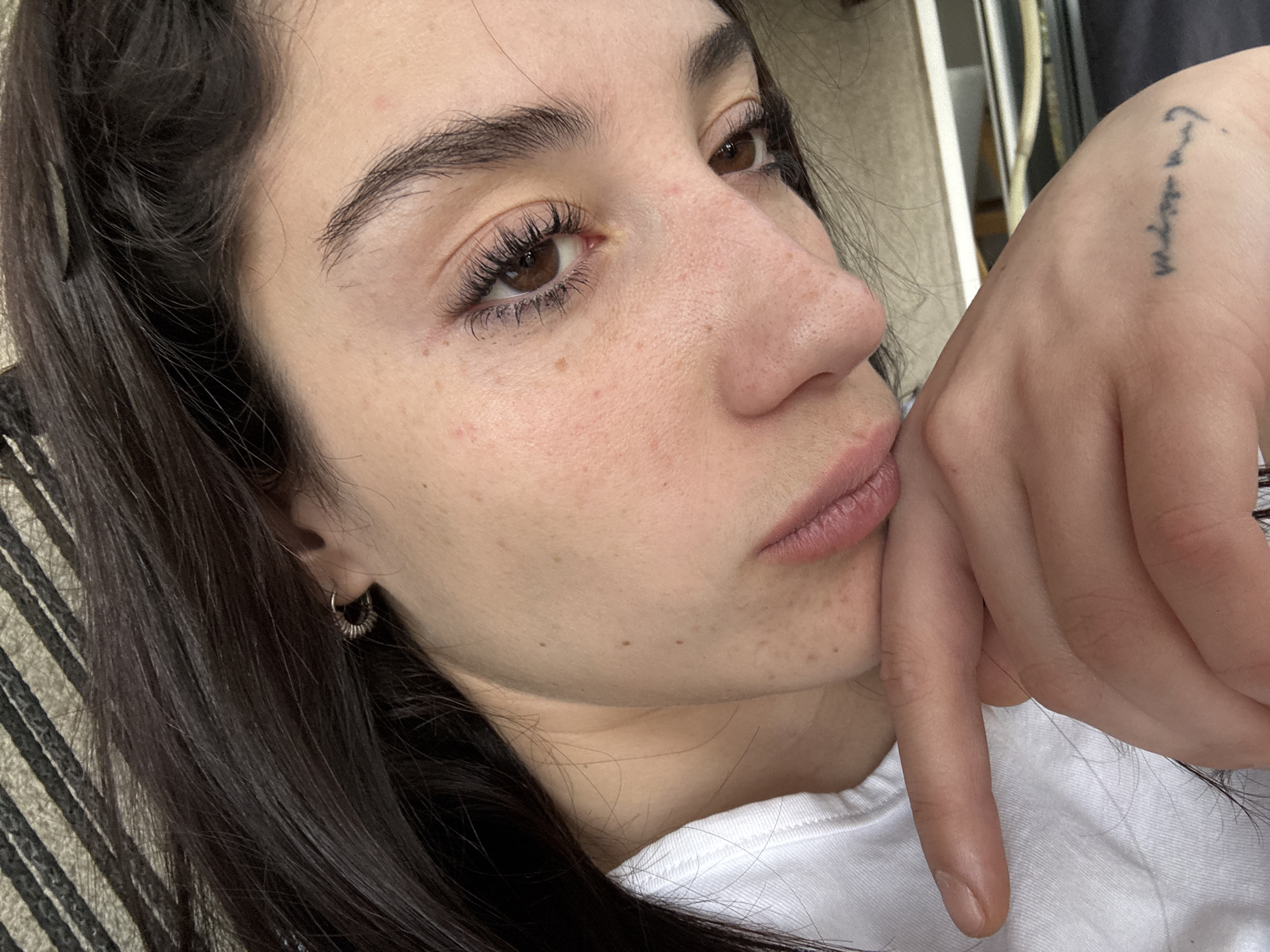 A close-up, low-angle shot focuses on a woman with long, dark hair and light skin. She has brown eyes and well-defined eyebrows. Her skin has some freckles. She is wearing small hoop earrings in her left ear. She has a tattoo on her right hand. Her finger touches her lips. She is wearing a white T-shirt. The background consists of neutral-toned blinds or panels. There is no indication of location, as there is nothing visible to suggest the city of Buenos Aires, Argentina.