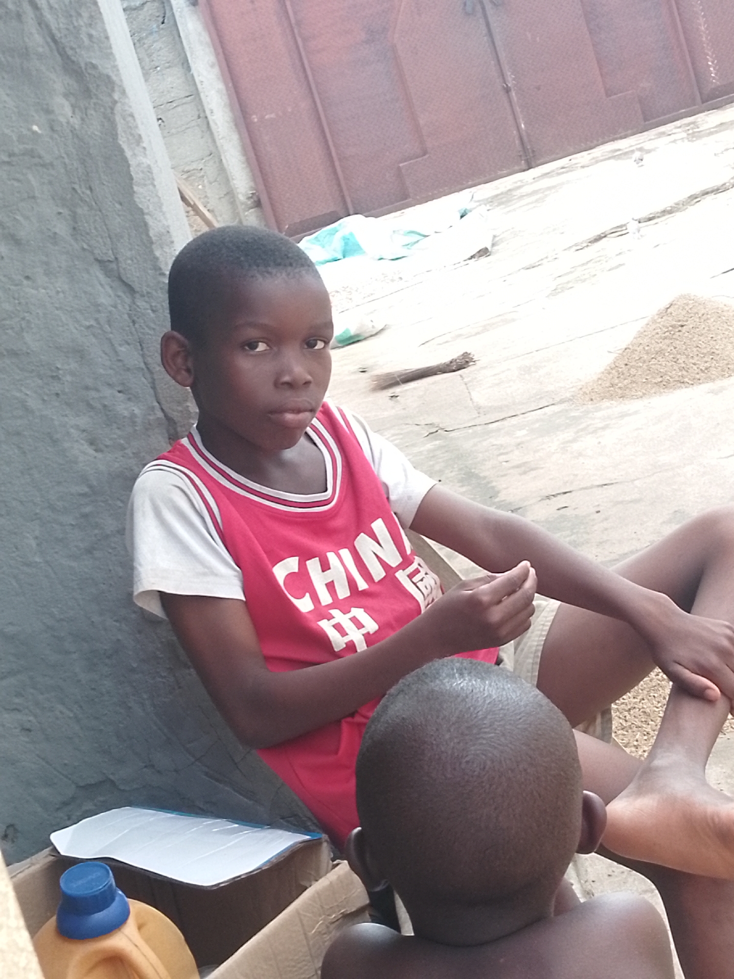 In this image, two young boys are the primary subjects. The boy in the foreground, seen from the back, has a closely shaved head and is wearing a dark t-shirt. He is seated facing away from the camera, towards the other boy. The second boy, more prominent in the image, is seated and looking directly at the camera. He is wearing a red jersey with white trim and a white t-shirt underneath. The red jersey has white text that appears to say "CHIN" and Chinese characters. He has light-colored shorts on.
The setting appears to be outdoors, possibly in a courtyard or an open area with a concrete floor. To the left of the boys, there is a textured gray wall. Behind them, a large, dark reddish-brown door or gate is visible, with a rough surface. The ground is scattered with what looks like loose grains or small stones. In the lower left corner, a cardboard box is partially visible, containing a yellow container with a blue cap and a white rectangular object, possibly a piece of paper or plastic.
The lighting suggests it might be daytime, with diffused light, possibly indicating an overcast day or shade from a nearby structure. The overall mood is casual and candid, with the boy in the red jersey presenting a serious expression. No specific activity or event is clearly depicted, other than the boys posing or resting in the described setting. The image is taken from a slightly high angle, looking down at the subjects.