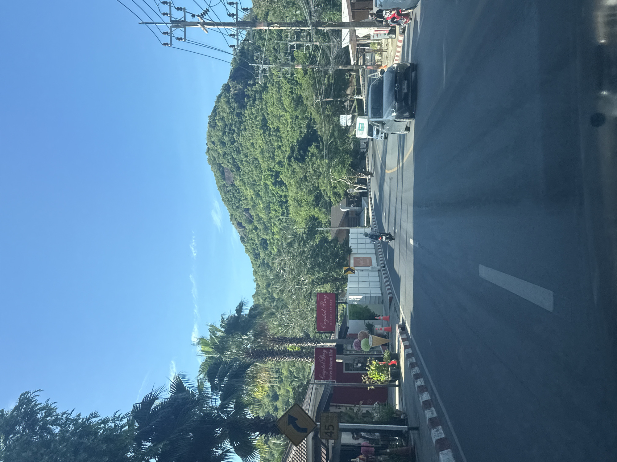 The image depicts a view from a vehicle moving along a paved road in Baan Chaweng Noi, Thailand. The road curves gently to the left, with a lush, green, tree-covered mountain dominating the background under a clear blue sky. To the left of the road, there are buildings with tiled roofs and signs, including one for "Crystal Bay" which appears to be a restaurant and bar, and another for "Crystal Bay Beach Resort". A large, colorful ice cream cone sculpture is displayed on the sidewalk. Several orange traffic cones are visible along the roadside.
Vehicles are present on the road: a dark-colored car is in the foreground to the right, a white van is further ahead, and a motorcycle with a rider is approaching from the opposite direction on the left side of the road. Another motorcycle, red in color, is parked on the right side of the road. There are power lines and utility poles lining the right side of the road. Palm trees are scattered throughout the scene, particularly on the left. The overall impression is a sunny day in a tropical, resort-oriented area.