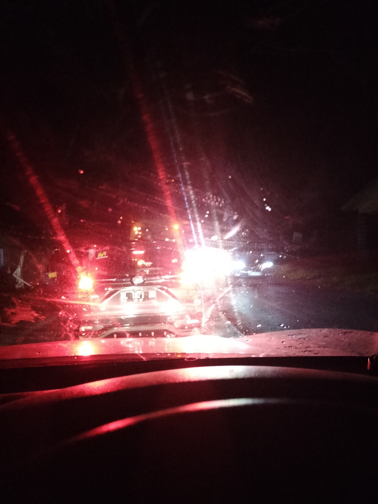 The image is taken from inside a vehicle at night, looking through the windshield. The windshield appears wet, with streaks and water droplets distorting the view. The scene outside is dominated by the bright headlights and taillights of other vehicles, creating lens flares and reflections. In the foreground, the dashboard of the vehicle is visible, with a reddish glow from the taillights of a vehicle directly ahead. Further ahead, there are multiple vehicles, their lights indicating a traffic situation. The overall impression is a view of traffic at night, possibly in a city or town, given the presence of streetlights and buildings faintly visible in the background. The location of Alue Dua Bakaran Batee, Indonesia, cannot be determined from the visual information in the image.