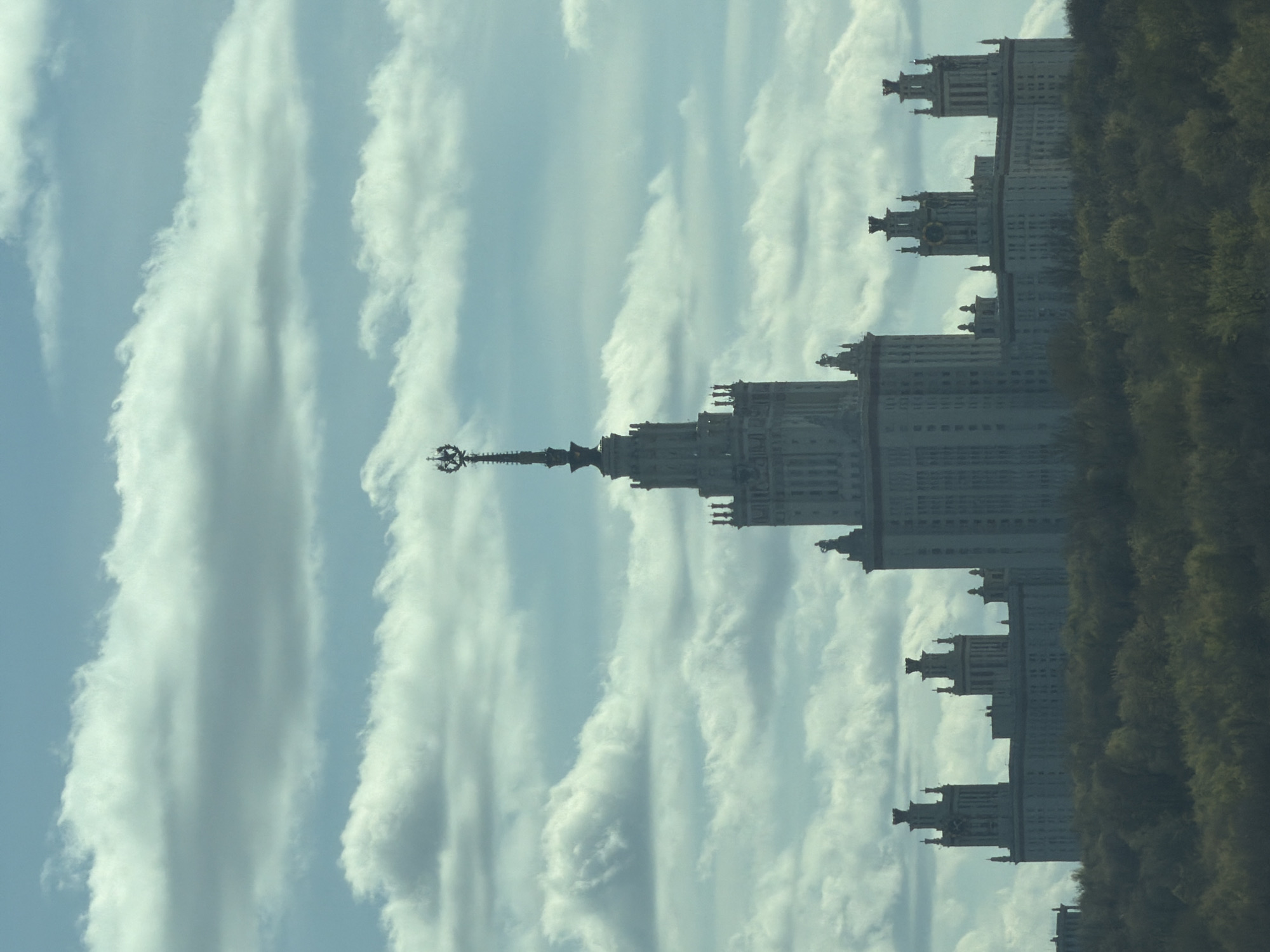 The image shows the Moscow State University building against a backdrop of cloudy skies and trees. The building is a Stalinist skyscraper with a prominent spire topped with a star. The location is Moscow, Russia.