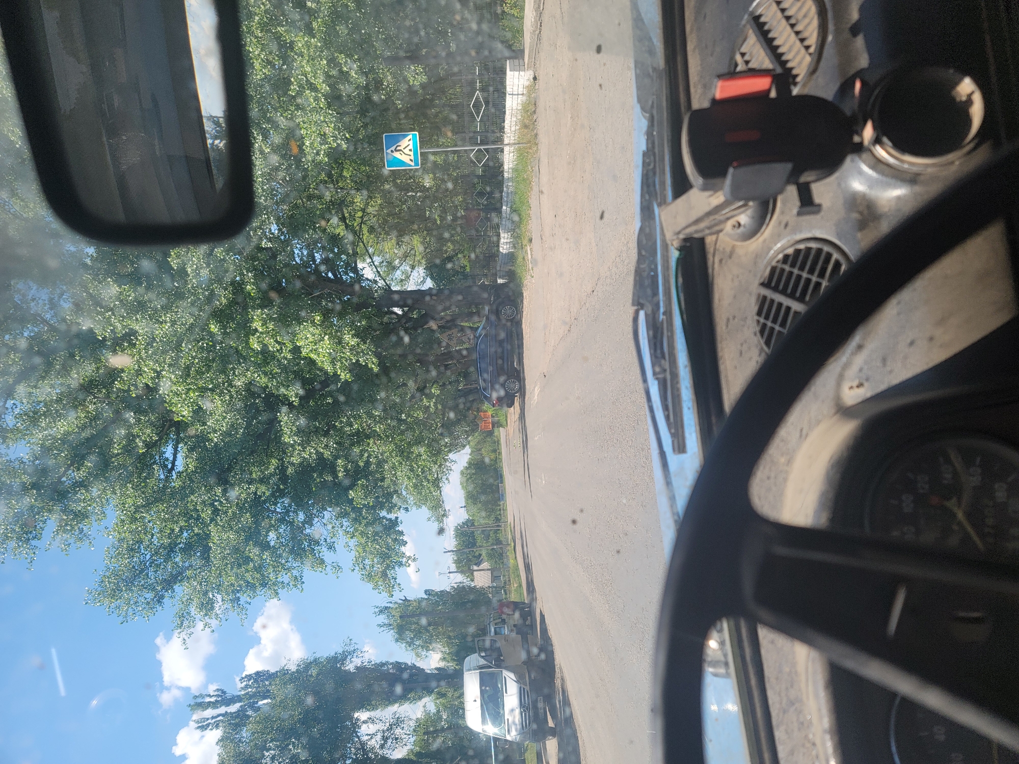 The image captures a street scene from the perspective of a driver in Pereshchepyne, Ukraine. A grey Mercedes-Benz Sprinter van with its side door open is parked on the left side of the road. Two individuals, one wearing a red shirt, are standing near the van. Further down the road, a black car is parked on the right side, near a pedestrian crossing sign. The road is paved and appears to be in a residential or suburban area, with trees lining both sides and houses visible in the background. The sky is blue with scattered clouds. The interior of the car shows the steering wheel, dashboard with a phone mount, and a speedometer.