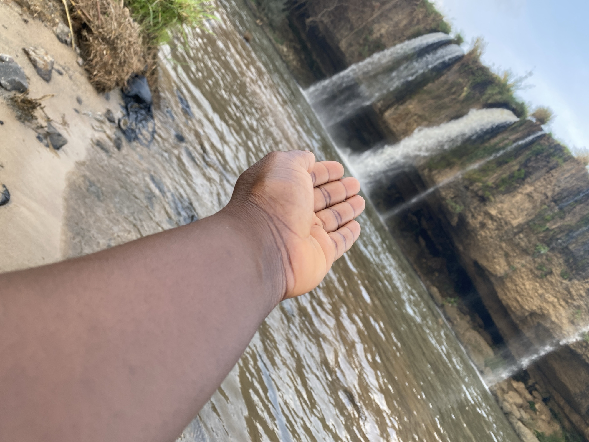 An eye-level shot shows a right human arm and hand extending out toward a waterfall, possibly somewhere in the city of Maigizo, Nigeria. The hand is brown-skinned and open. The arm is in the lower left portion of the frame. The waterfall is in the background. The water is flowing over a rocky cliff face. There is vegetation on the cliff face near the waterfall. The sky is visible in the upper right portion of the frame. In the lower left of the frame there is a small beach with rocks and debris on it.