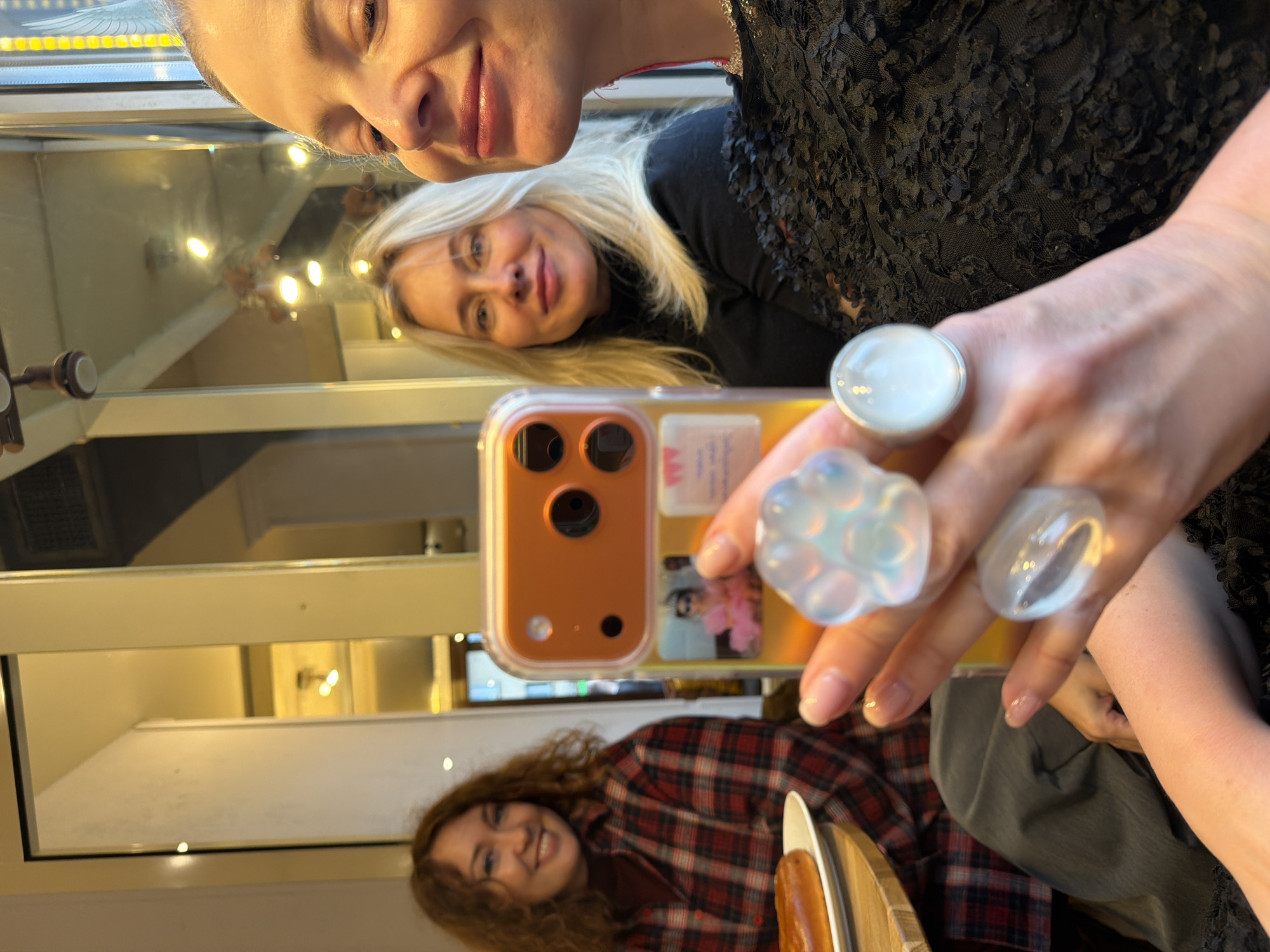 Three women are visible, apparently taking a selfie in front of a mirror. The mirror's reflection shows an interior space. One woman with blonde hair is wearing a black, textured top, and holding a cell phone with an orange case in front of her. She has large rings with bulbous designs on her fingers. Two other women are reflected in the mirror; one with blonde hair and one with curly brown hair wearing a plaid shirt. The location is identified as Moscow, Russia.
