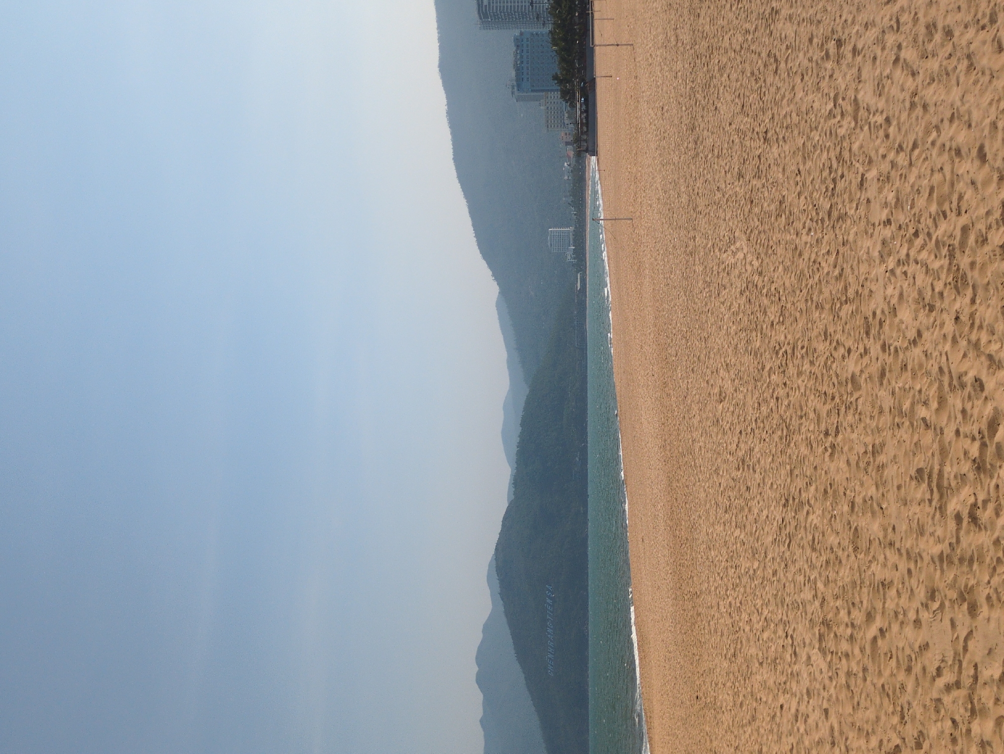 The image depicts a beach scene, likely in Quy Nhon Ward, Vietnam. The beach is expansive, covered in golden-brown sand marked with footprints. To the left, a line of greenish-blue water meets the shoreline. Further inland, a mountain range rises, dotted with vegetation. Buildings are visible near the mountains. Above, a pale, hazy sky fills the frame.