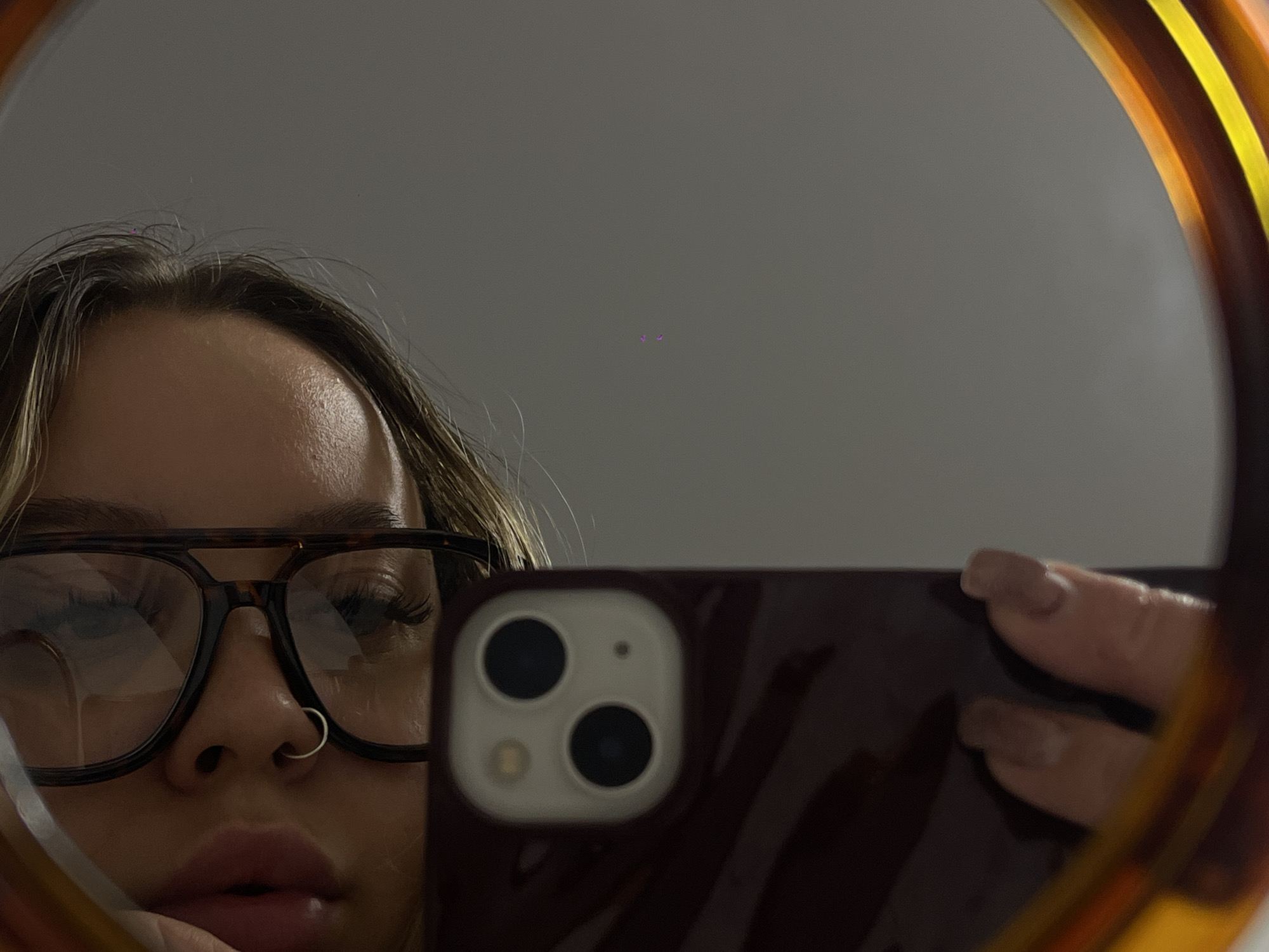 This media file captures a mirror selfie taken in Krasnodar, Russia, focusing on the reflection of a person from the forehead to the chin. The image is framed by a distinct tortoiseshell-patterned mirror border. The background reflected in the mirror is a plain, light grey wall, with a small purple anomaly in the upper center.
The subject, likely female, has light-colored hair partially visible at the top. They are wearing dark, tortoiseshell-patterned eyeglasses, reflecting parts of the room and the phone. A silver hoop nose ring is prominent on their right nostril. The person's lips are full, and their eyes, adorned with long eyelashes, appear to be looking downwards.
The person is holding a smartphone with a dark red or maroon case, which obscures much of the lower right portion of the face. The phone's dual camera module is clearly visible on its back. The subject's hand and fingers, with light-colored fingernails, can be seen holding the device. The lighting is soft and indirect, creating a slightly subdued atmosphere. There is no visible text in the image.