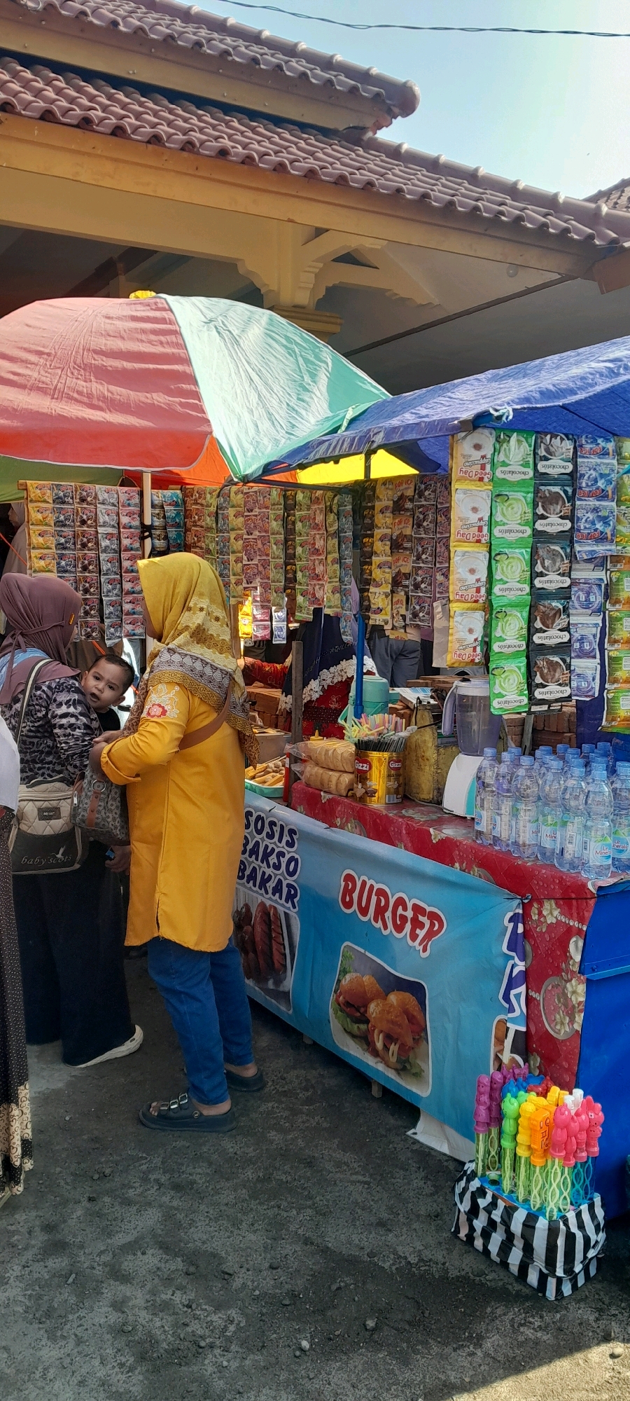 The image captures a street-side food stall under a colorful awning in Jeruklegi Kulon, Indonesia. The stall appears to be selling snacks, drinks, and potentially grilled items. In the foreground, several bottles of water are neatly arranged on the counter, alongside a display of packaged food items hanging from racks. A blue banner at the front of the stall features images of burgers and text that reads "BURGER?" and "SOSIS BAKSO BAKAR," indicating the types of food available. To the left, a woman in a bright yellow headscarf and matching tunic, with blue jeans, is interacting with the stall. A child with wide eyes peeks out from behind another person, also wearing a headscarf and a patterned top, positioned to the left of the woman in yellow. The ground is a rough, gray surface, likely asphalt or concrete, with some scattered debris. In the background, a tiled roof of a building is visible, suggesting a semi-urban or market setting. To the right, a collection of colorful bubble wands in a striped container adds a playful element to the scene.