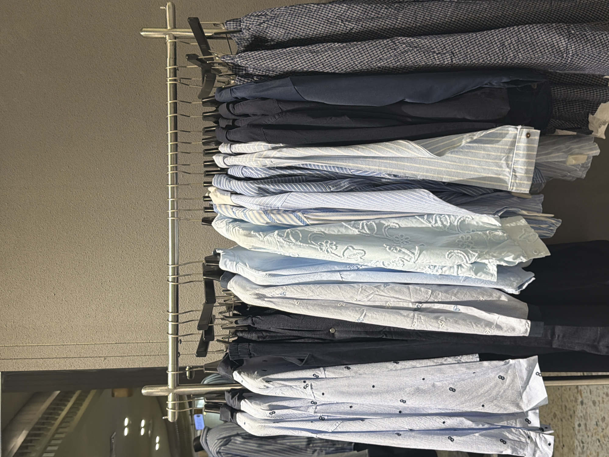 A row of light blue and dark blue men's shirts are hanging on a metal rack in what appears to be a clothing store in East Jakarta, Indonesia. The shirts are a variety of patterns and solid colors, with some featuring subtle designs like stripes or small dots. The background is a plain, textured grey wall. The lighting suggests an indoor environment.