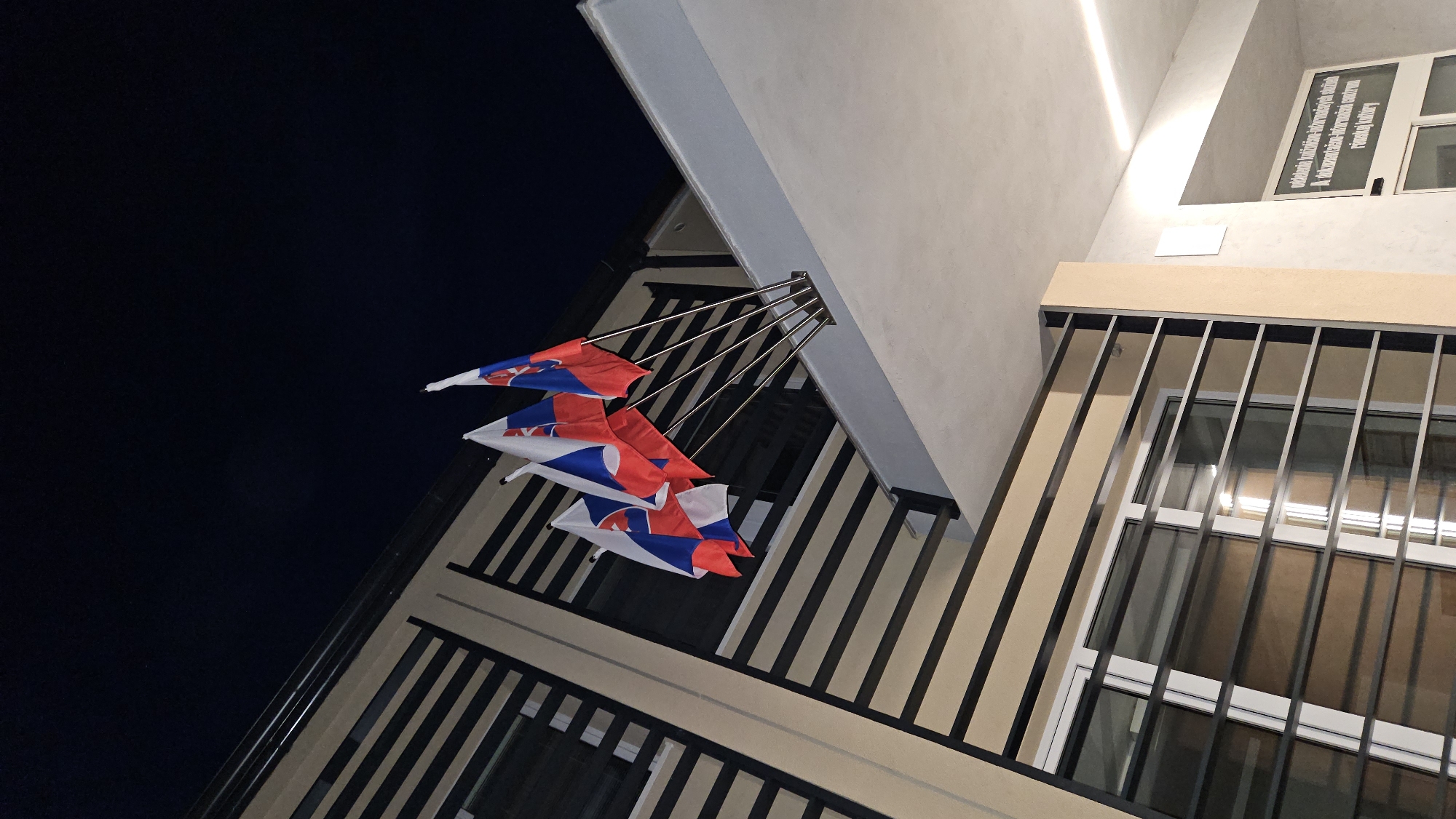 Four Slovak flags are displayed on a building's exterior at night. The flags are mounted on metallic poles extending from a bracket on a light-colored overhang. The flags are red, white, and blue with the Slovak coat of arms. The building has a modern architectural design with horizontal beige and dark-colored louvers over windows. The sky is dark blue, indicating nighttime. A sign in Slovak is visible on the building to the right, which reads "oddelenie knižnično-informačných služieb" and "A dokumentačno-informačným centrom rímskej kultúry". This signage suggests the building may be a library or cultural center. Based on the signage and the flags, the location is likely the Jonáš Záborský Library in Prešov, Slovakia, which houses a documentation and information center for Roman culture. There are no people visible in the image.