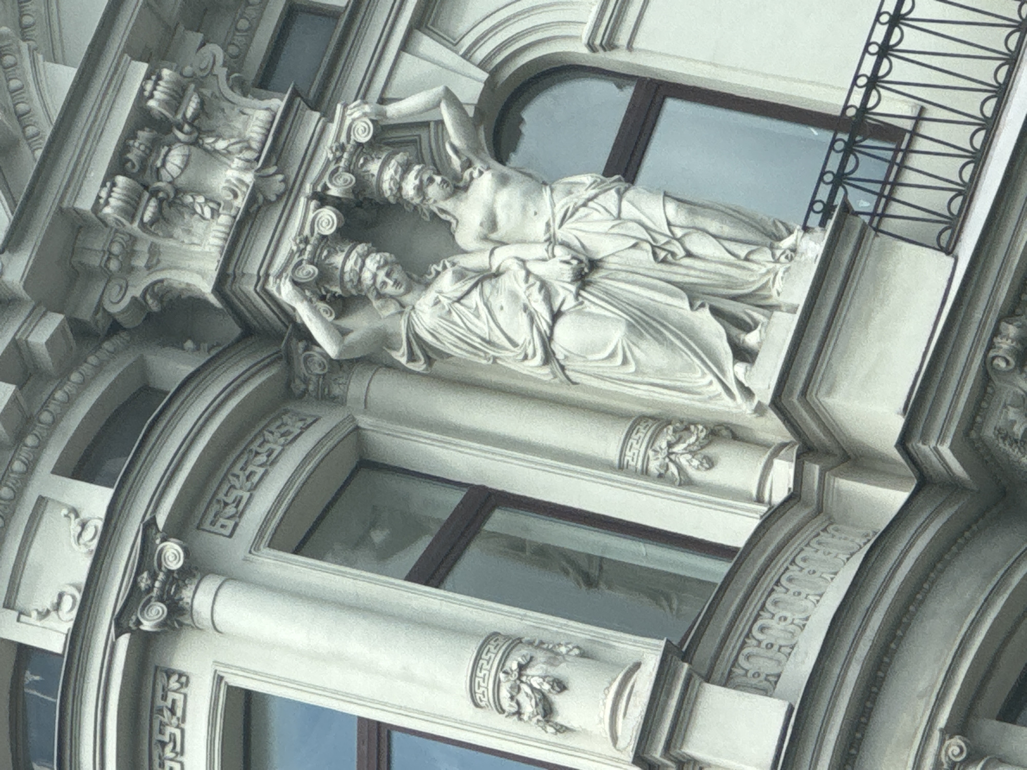 The image shows architectural details of a building, likely taken in Saint Petersburg, Russia. The focus is on the façade adorned with decorative elements, specifically two reclining female statues above windows. The statues are classical in style, draped in fabric. Above the statues, there is decorative crown-like headwear. The building's exterior is light-colored. A decorative metalwork detail is visible at the window.