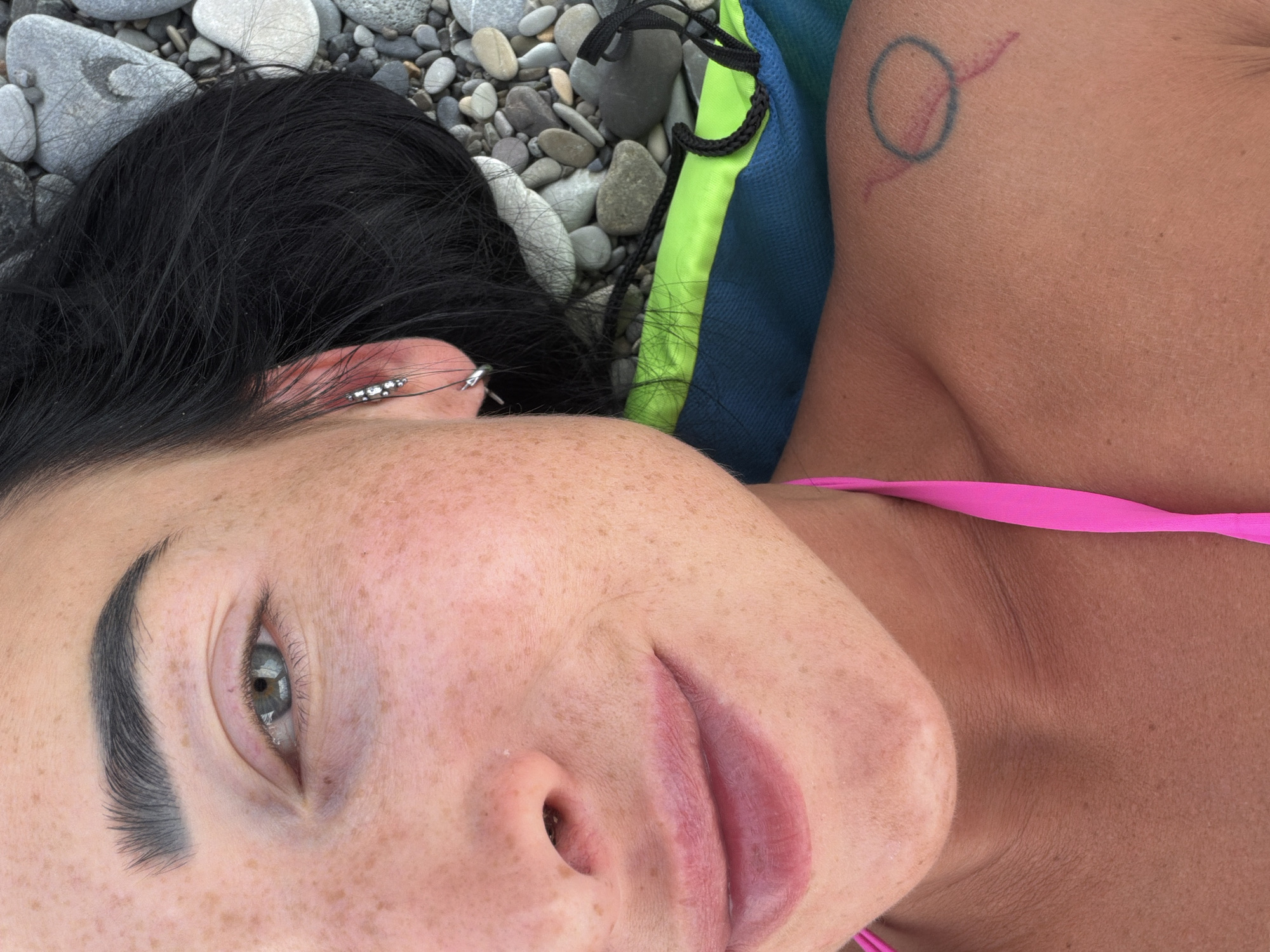 This media file is a close-up, high-angle shot of a person lying on a pebbled surface, possibly a beach. The person has dark hair and is wearing a pink bikini top. Their face is partially visible, with freckles scattered across their skin. They have a tattoo on their shoulder, a blue circle with a pink line extending from it. The background consists of grey and white pebbles, with a glimpse of a blue and green fabric, possibly a towel or bag. The lighting suggests it is daytime. The overall impression is one of a relaxed, outdoor setting.