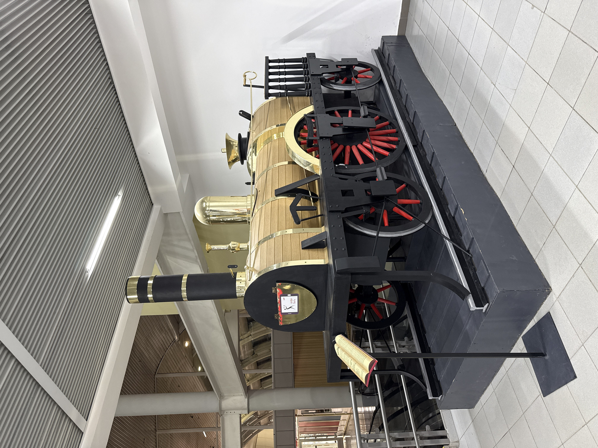 This is a media file of a decorative, life-sized model of an old steam locomotive. The locomotive is made of wood and brass, with black metal wheels that have red spokes. It is displayed indoors, likely in a museum or public space, given the white tiled floor and corrugated metal ceiling.
The locomotive is the central object in the scene. It is positioned on a dark platform, which is itself on the tiled floor. The details of the locomotive are intricate, including a large smokestack, a boiler, and exposed mechanisms. There are also some decorative elements, such as a golden bell and a handle near the top of the boiler. A small plaque with text is visible on the side of the locomotive, and an open book with red pages is resting on a railing in front of it.
The setting appears to be a modern building with a high ceiling and large windows or skylights that provide ample lighting. The background shows structural elements of the building, including beams and more of the corrugated ceiling. The overall impression is one of a static display, possibly an exhibit. There are no people visible in the frame, and no indication of the time of day or weather conditions. The location cue "Novokuybyshevsk, Russia" suggests this is the geographical context of the image. The visible text on the plaque is too small to be legible in the provided image.