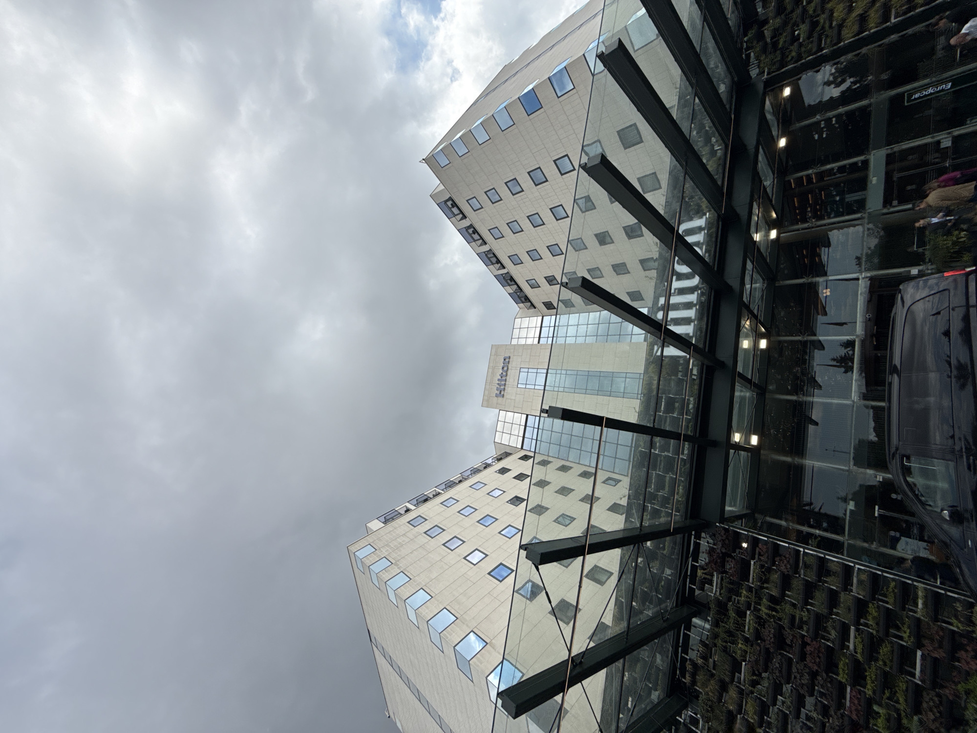 The image presents an upward-angled view of modern architecture in Sofia, Bulgaria, under an overcast sky. Two large, contemporary buildings dominate the scene. One building, centrally located and slightly further back, is identified by the "Hilton" logo visible on its glass and light-colored facade. This building features a pattern of small, square windows and larger glass sections.
In the foreground, a prominent glass and metal canopy or structure extends, providing multiple reflections of the surrounding environment. This reflective surface mirrors parts of the buildings, the cloudy sky, and elements at ground level. Among these reflections, the word "Europcar" is discernible, alongside partial views of people and a dark-colored vehicle, possibly a van or SUV. Adjacent to or part of the structure in the foreground, a distinctive vertical green wall or garden feature is visible, adding a natural element to the urban setting.
The overall impression is that of a bustling urban area, likely the entrance to a hotel or a commercial complex. The cloudy weather suggests a daytime shot with diffused light, contributing to the muted color palette of the sky and the building facades.