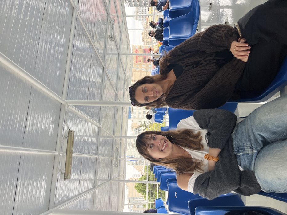 Two adult females are seated on a passenger ferry deck in Piraeus, Greece. The deck is covered by a white metal framework supporting a translucent, ribbed roof, with fluorescent light fixtures attached. Rows of bright blue plastic bucket seats are present, with some occupied by other passengers in the background.
The woman on the left, positioned closer to the camera, has long brown hair with bangs, wears a white off-the-shoulder top, a dark gray cardigan, and light blue denim jeans. She has gold hoop earrings and an orange scrunchie on her right wrist, and smiles directly at the camera with her arms crossed.
The woman on the right has long brown hair with sunglasses resting on her head. She wears a dark brown chunky knit cardigan over a black top and black trousers. Rings are visible on her fingers, and she holds a dark object in her lap. She smiles subtly towards the camera.
Beyond the ferry's side railing, an urban landscape of multi-story buildings and some trees is visible under an overcast sky, characteristic of a port city.