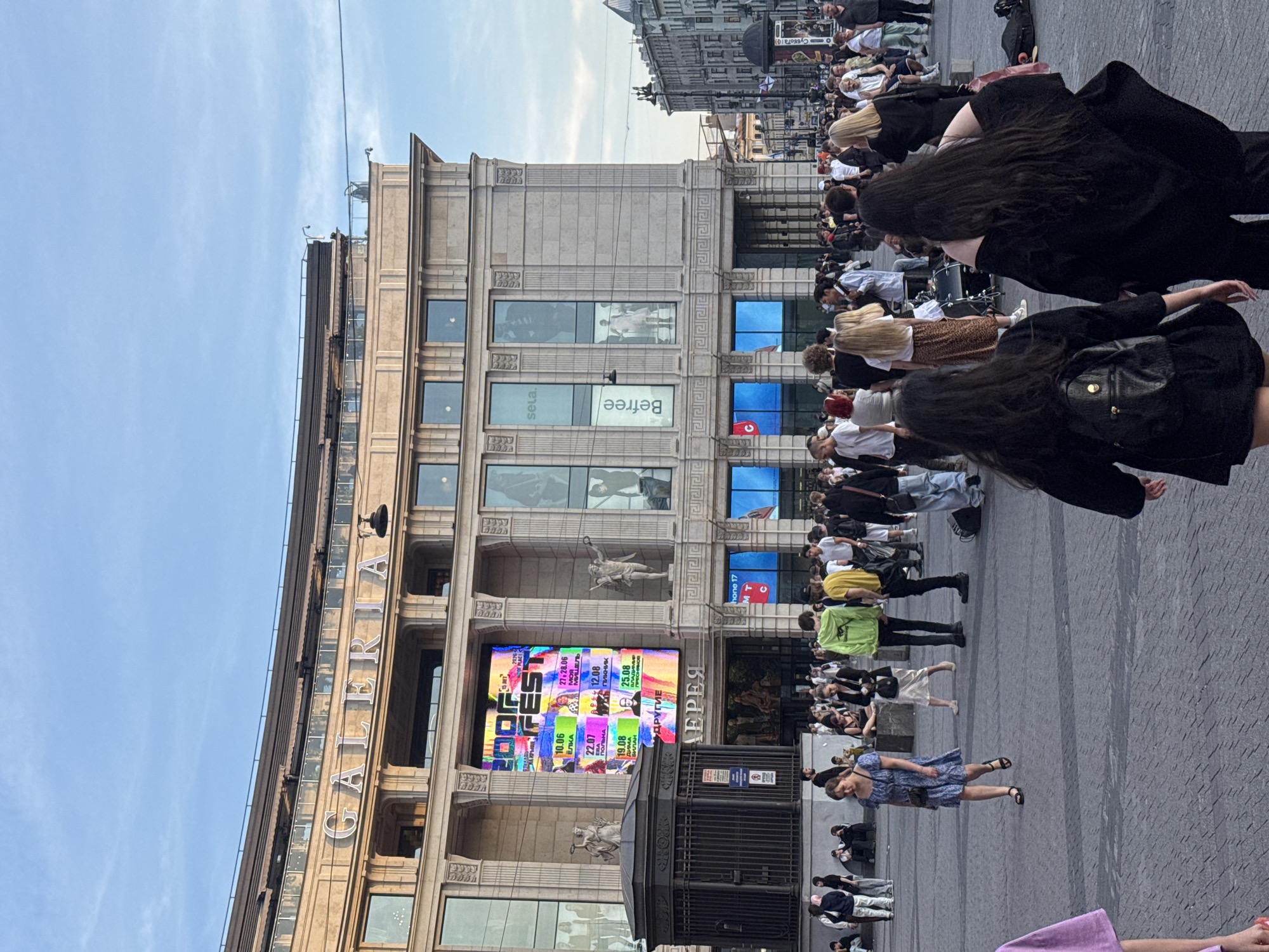 A low-angle, full shot captures a bustling urban square in front of a grand, multi-story building with the name "GALERIA" prominently displayed in large, illuminated letters across its facade. The building exhibits classical architectural elements, including columns and ornate detailing. A large digital billboard displays colorful graphics and text, featuring event listings with dates and times. Below the billboard, a dark, cage-like structure with intricate metalwork stands. The paved ground is populated by a diverse crowd of people, many walking or standing, while others are seated. Several individuals in the foreground are turned away from the camera, obscuring their faces. The sky above is a clear blue with soft clouds. Streetlights and architectural features of surrounding buildings are visible in the background, suggesting a city environment.